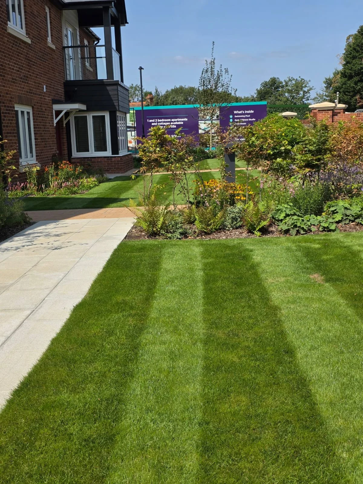 A well-maintained lawn with a shadow of a person taking the photo. There is a paved walkway on the left and garden beds with various plants and flowers. In the background, there's a multi-story brick building and a colorful sign with information abou