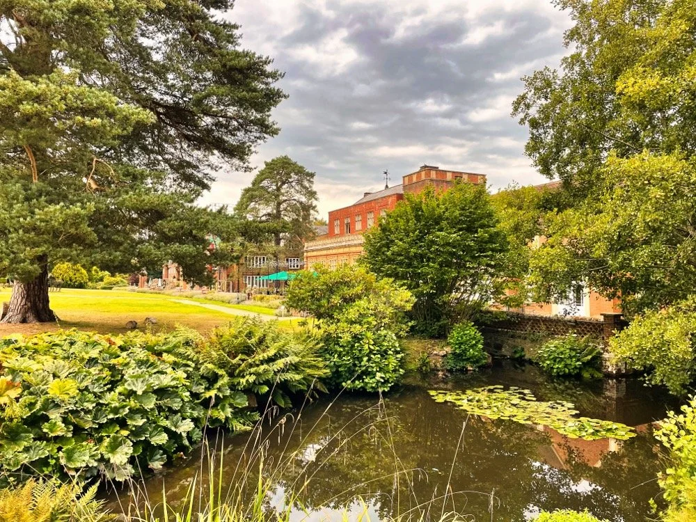 Scenic view of a park with a pond, surrounded by lush green trees and bushes, with a brick building in the background under a cloudy sky.