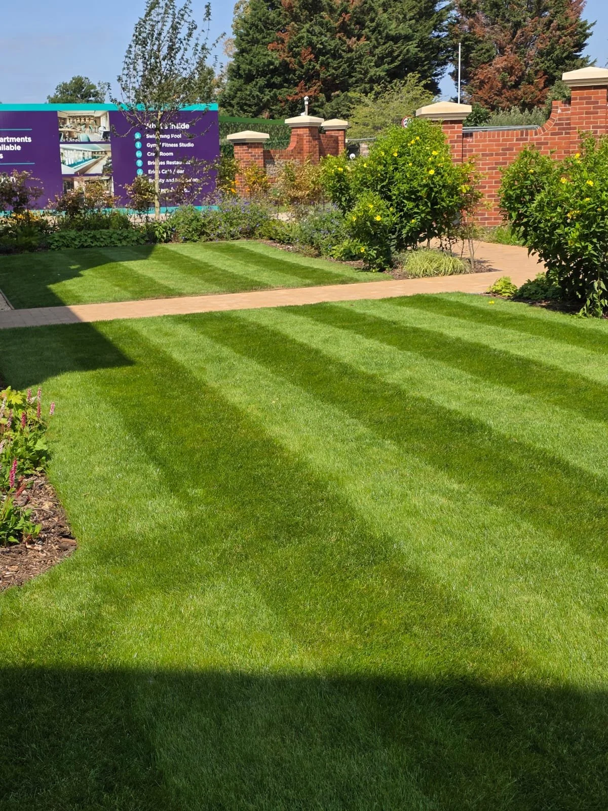 Well-maintained green lawn with light and dark striped patterns, bordered by flower beds and bushes, with a blue informational sign in the background, brick wall, and trees.
