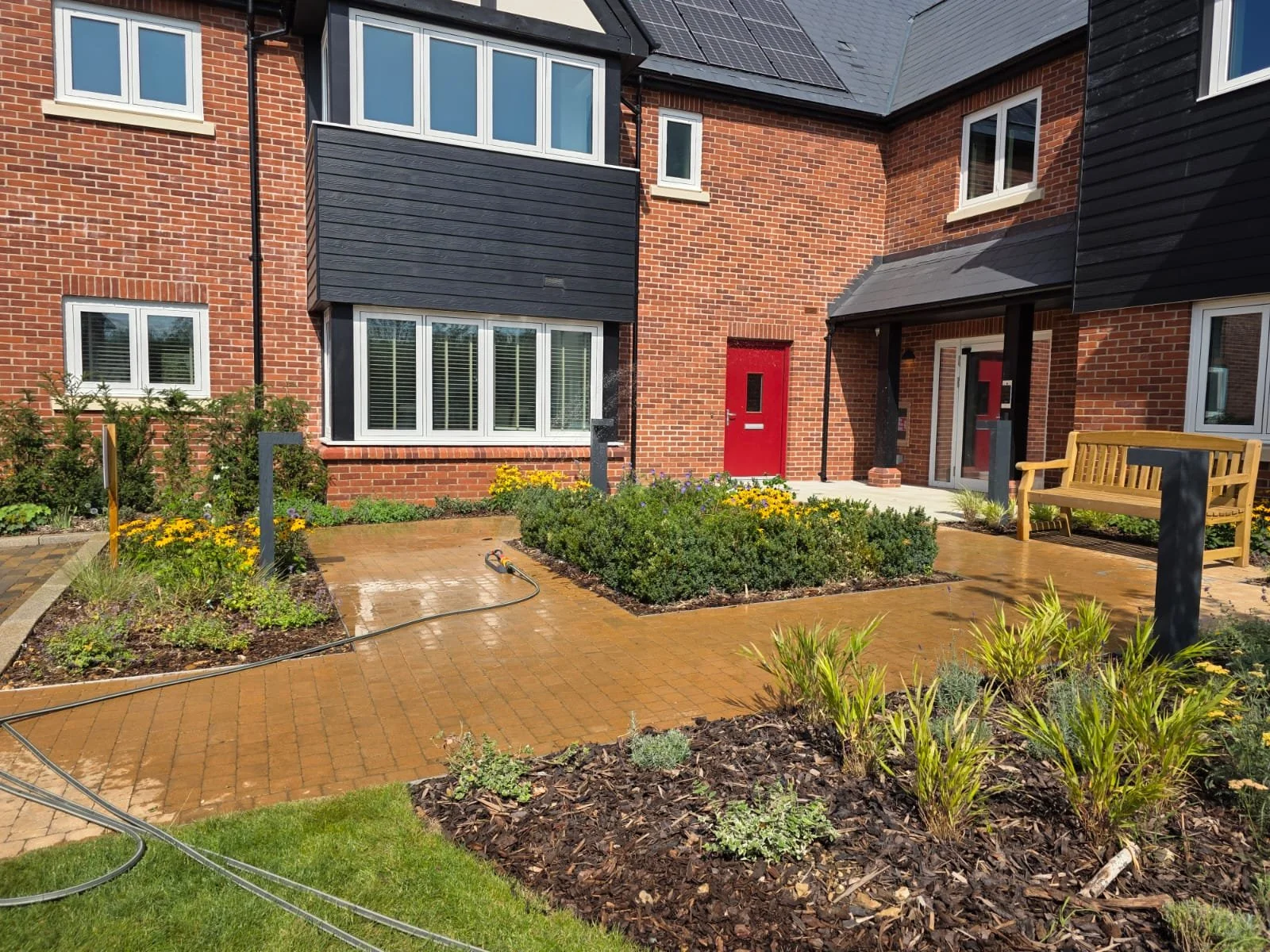 A view of an apartment complex courtyard with a red door, a black and brick exterior, landscaped flower beds, and a garden hose on a brown brick patio.