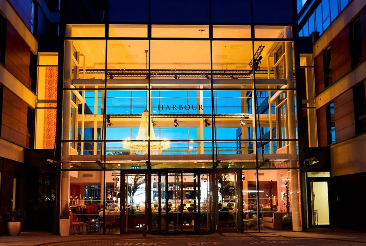 The entrance of a building named 'Harbour' with a glass facade, showing an interior illuminated with warm lighting, and a large chandelier visible inside.