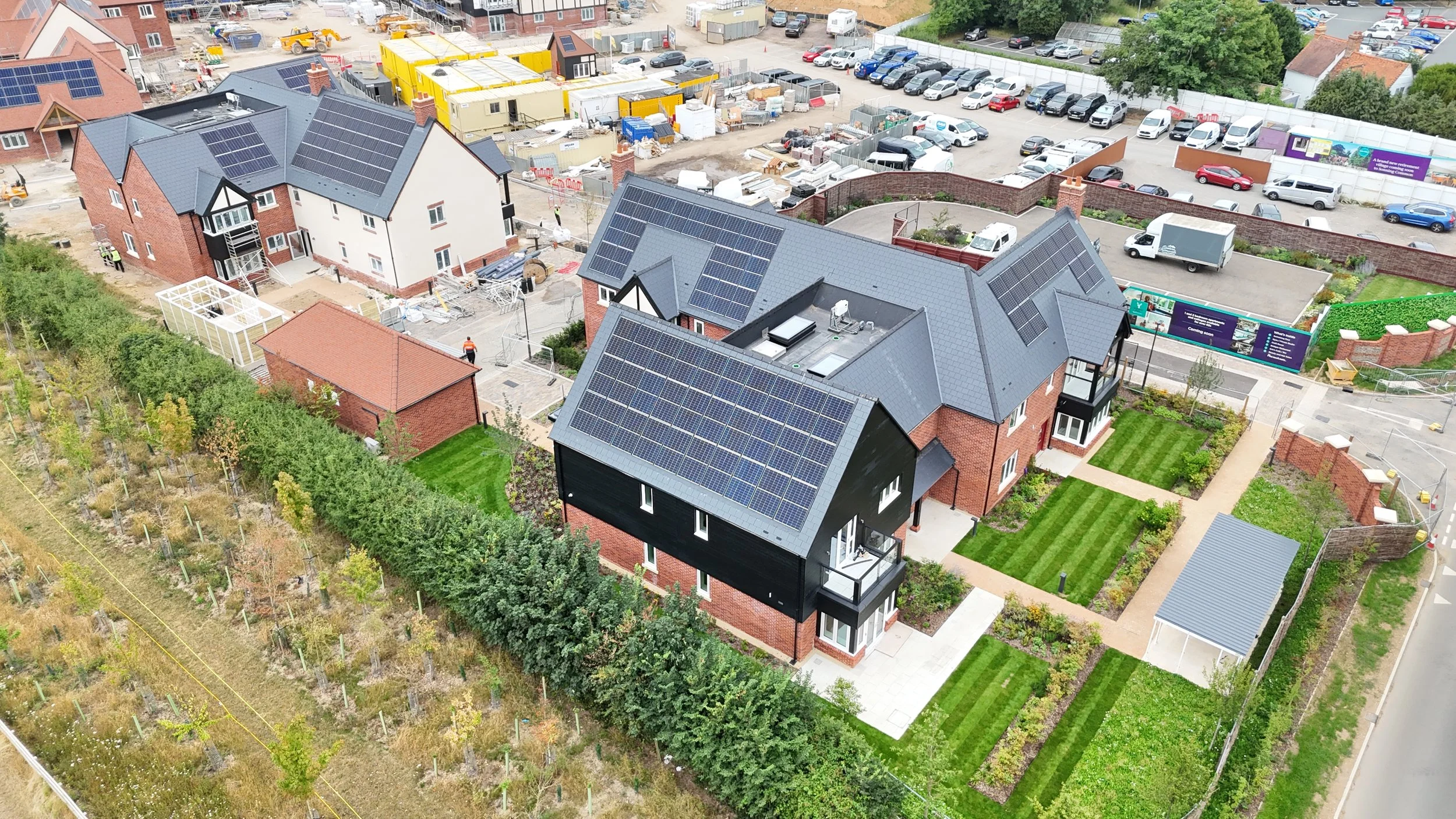 Aerial view of a residential area with modern houses featuring solar panels on roofs, surrounded by well-maintained lawns and gardens, with a construction site and parked cars in the background.