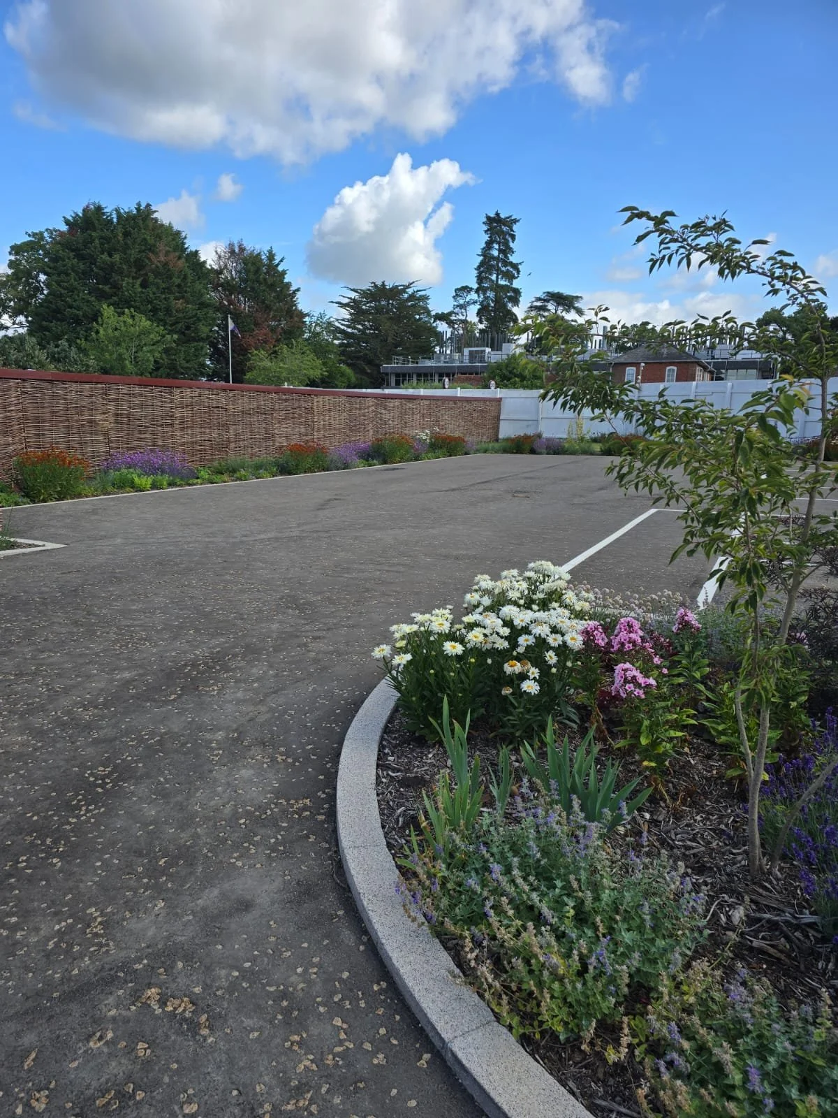 A parking lot bordered by colorful flowers and a low brick wall, with trees and buildings in the background and a partly cloudy sky.