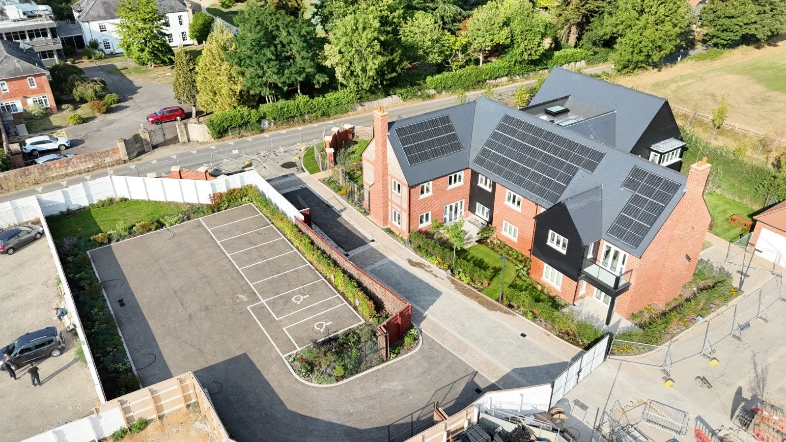 An aerial view of a residential area showing a brick apartment building with solar panels on the roof, a parking lot with designated handicapped spaces, and surrounding greenery.