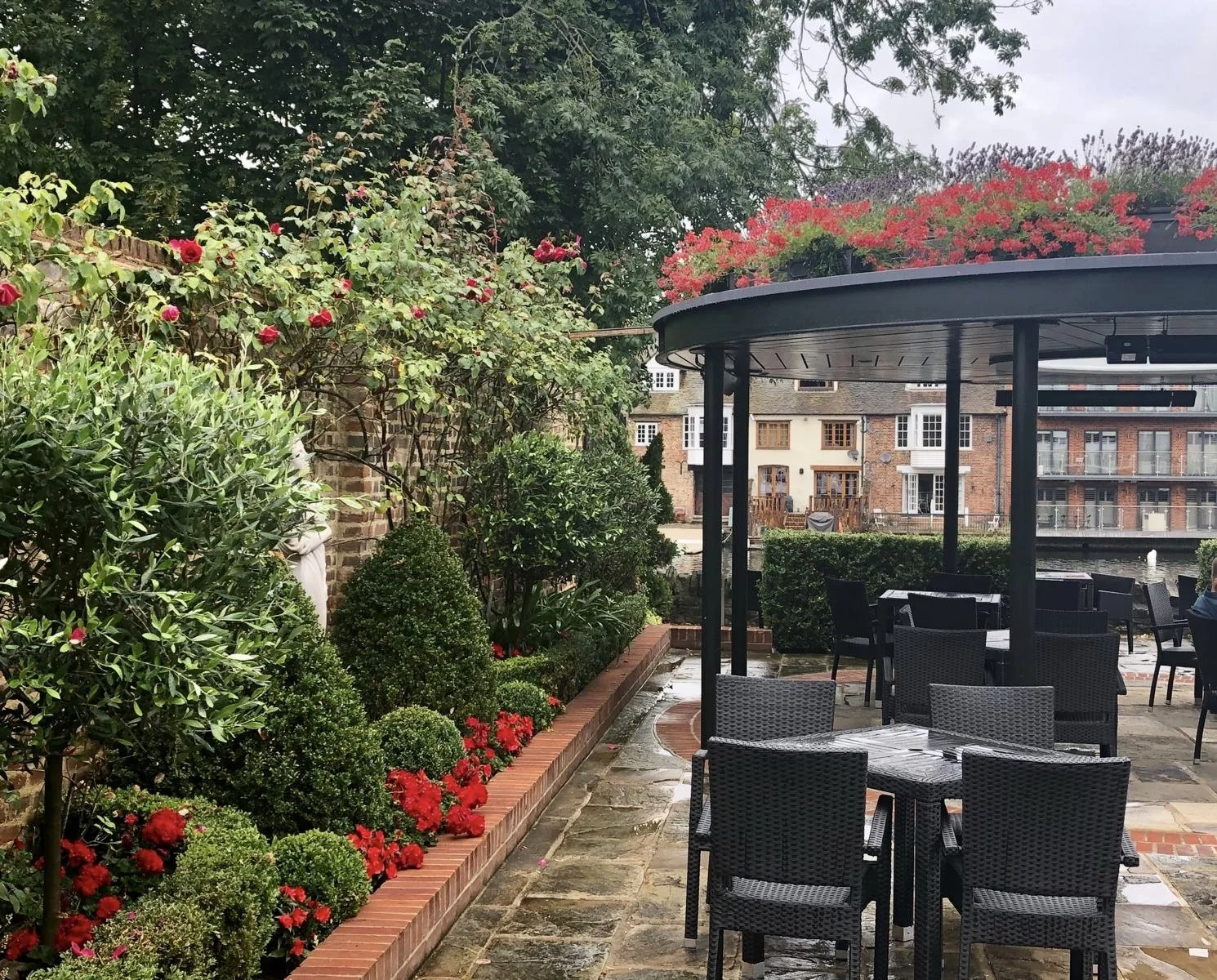Outdoor patio area with black wicker chairs and tables, a landscaped garden with bushes, flowering plants, and rose bushes along a brick border, surrounded by buildings and trees, on a cloudy day.