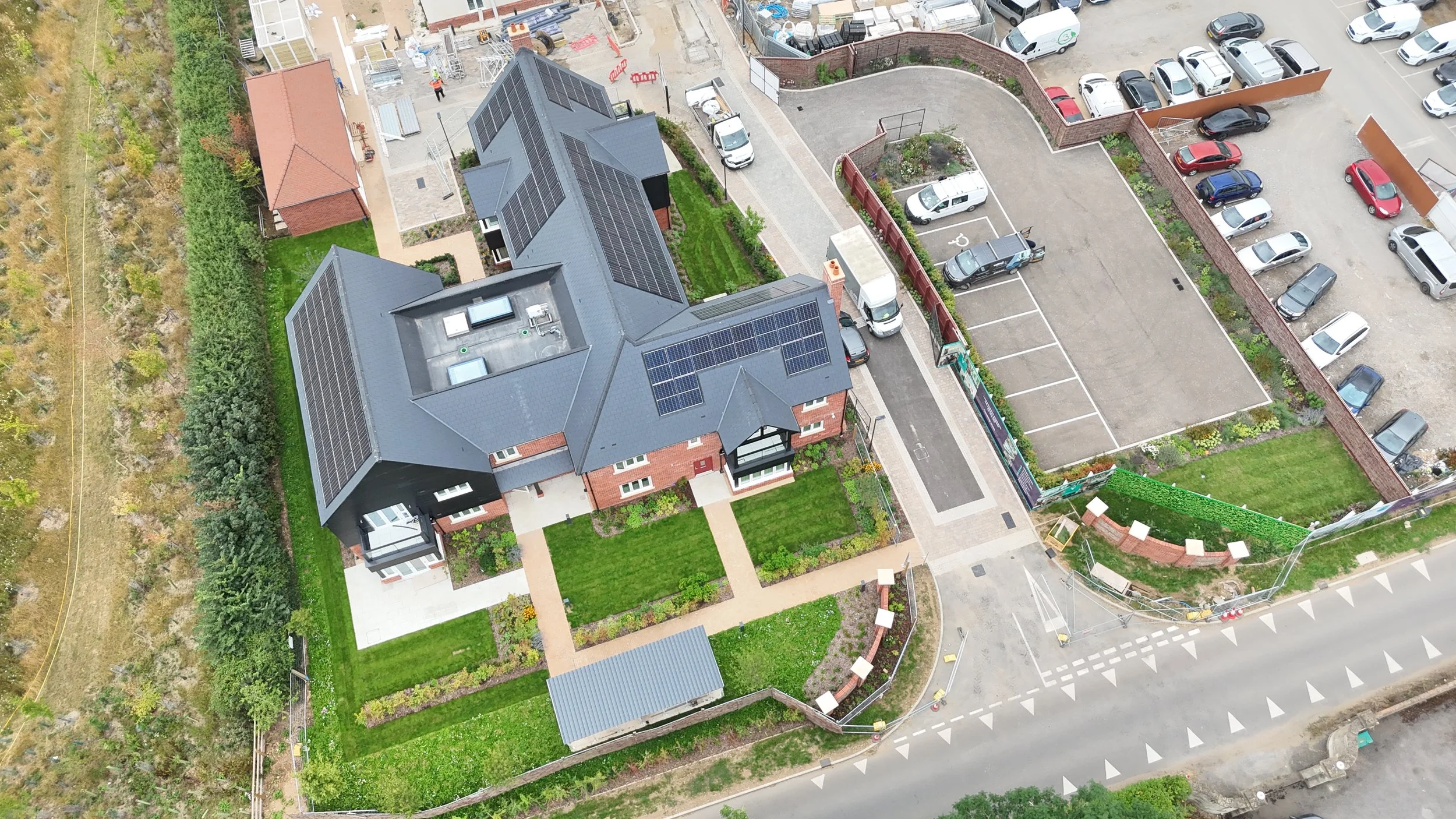 Aerial view of a modern residential house with solar panels on the roof, green lawns, a parking lot with several cars, and neighboring construction sites.