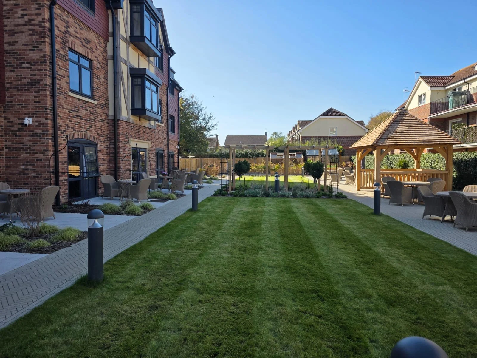 A landscaped outdoor patio area with a central grass lawn, surrounded by paved walkways, outside a brick residential building. There are outdoor seating arrangements, including chairs and tables, and a wooden gazebo in the background under a clear blue sky.