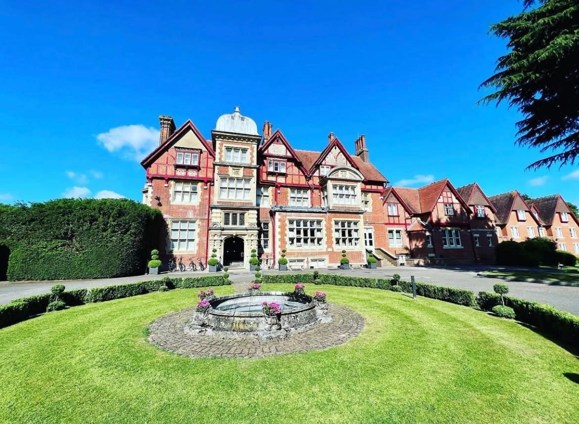 A large, historic mansion with a red brick and stone facade, multiple gabled roofs, and an ornate entrance, surrounded by well-manicured lawns and gardens, under a clear blue sky.