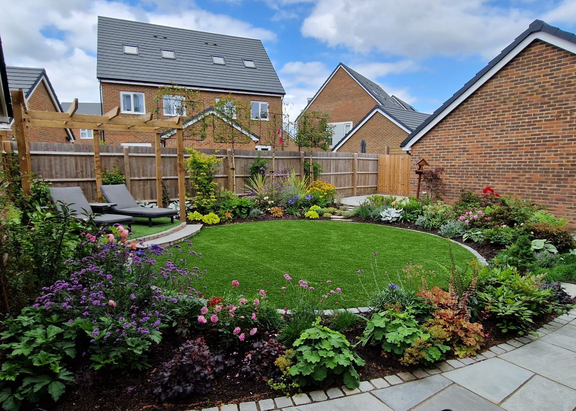 A well-maintained backyard garden with a green lawn, flower beds, and two lounge chairs under a wooden pergola. There are neighboring houses with brick walls and gray rooftops in the background under a partly cloudy sky.