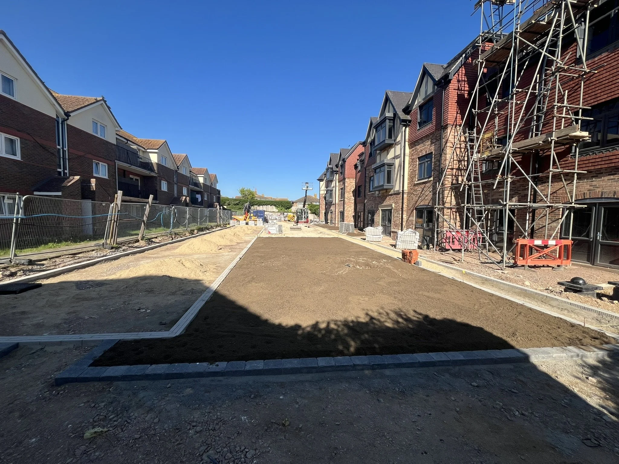 Under construction pathway with dirt and paving, scaffolding on right side, residential buildings on both sides, clear blue sky.
