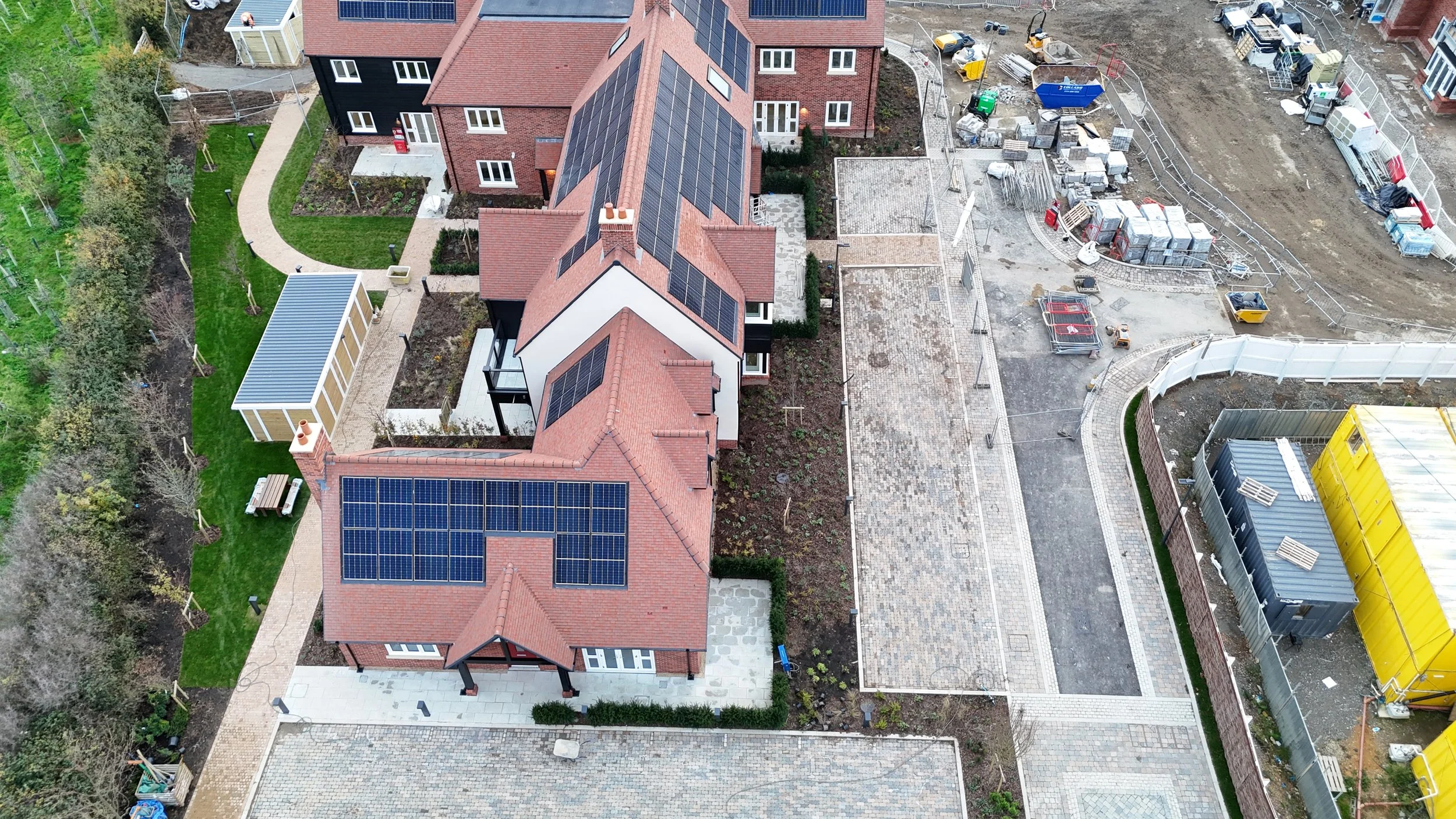 Aerial view of a residential area with new construction, showing houses with solar panels on the roofs, a landscaped yard, a garden shed, and a construction site with pavement work and building materials.