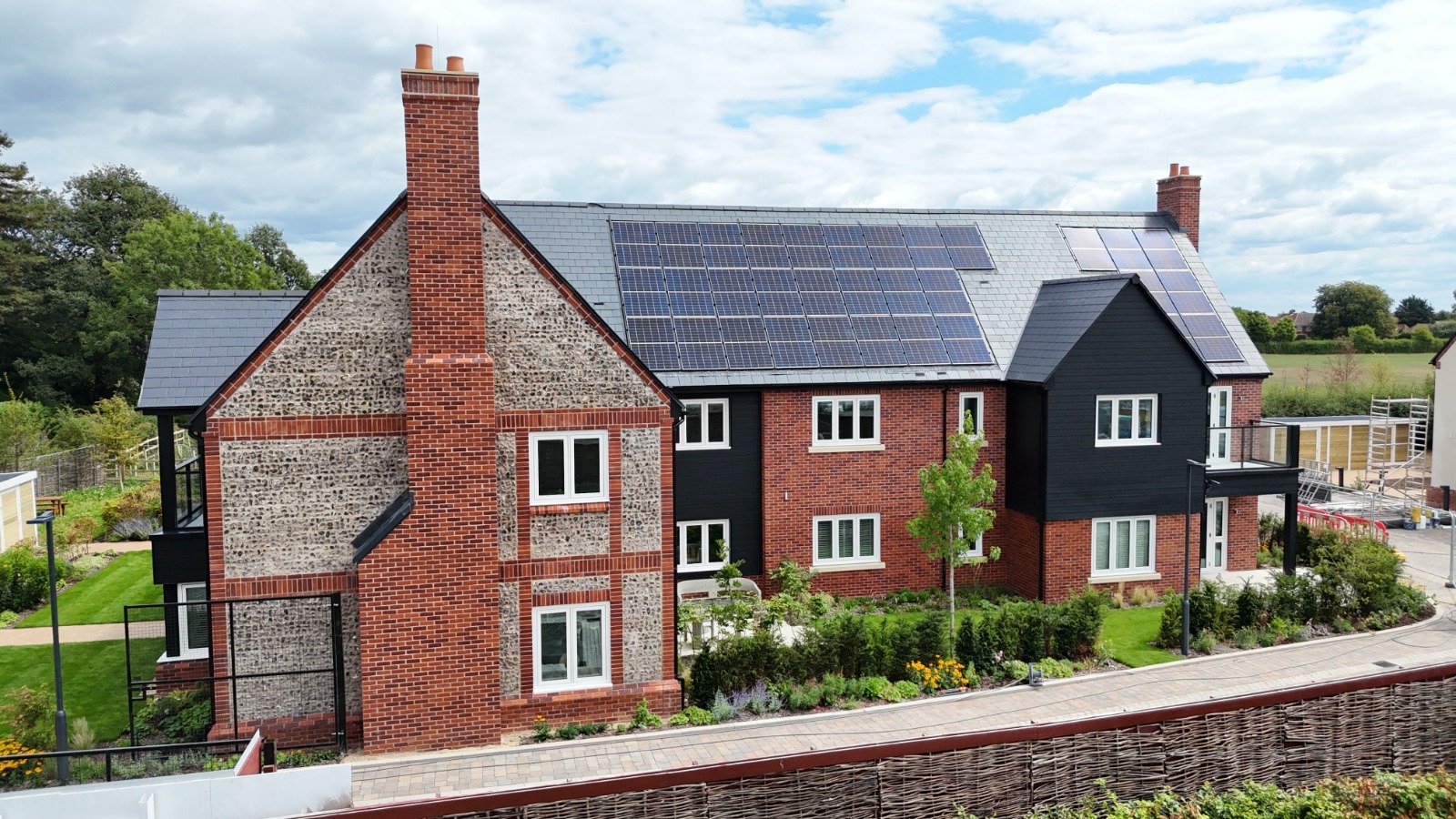 A house with brick and black exterior walls, equipped with solar panels on the roof, a courtyard with greenery and small trees, and a paved walkway in front.