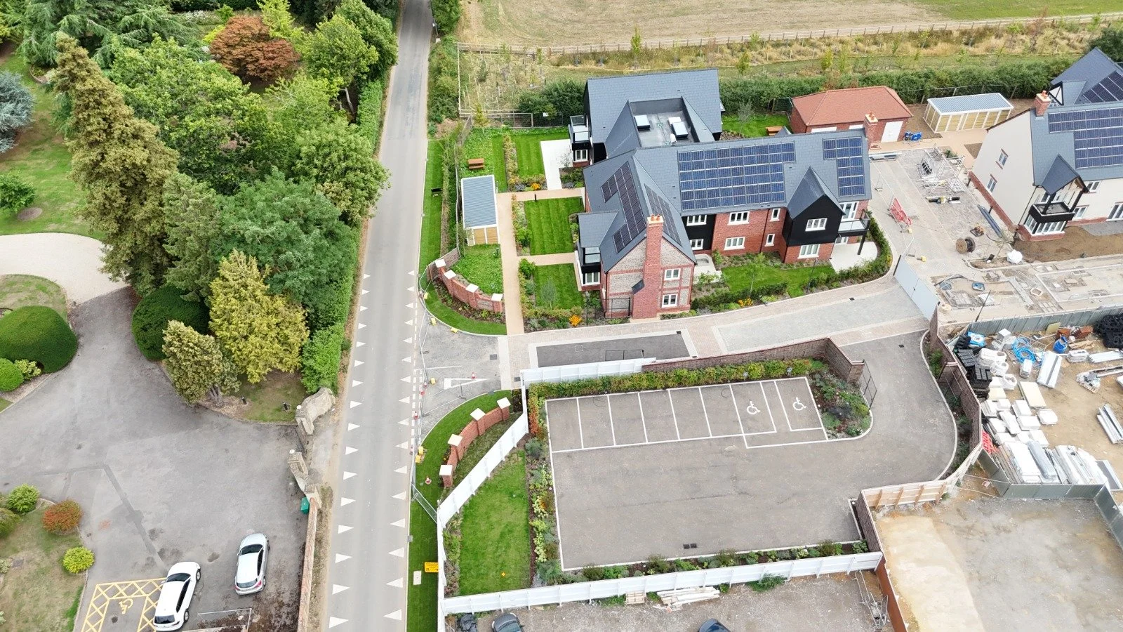 An aerial view of a residential neighborhood with modern houses, a parking lot with two handicapped spaces, green lawns, trees, and ongoing construction.
