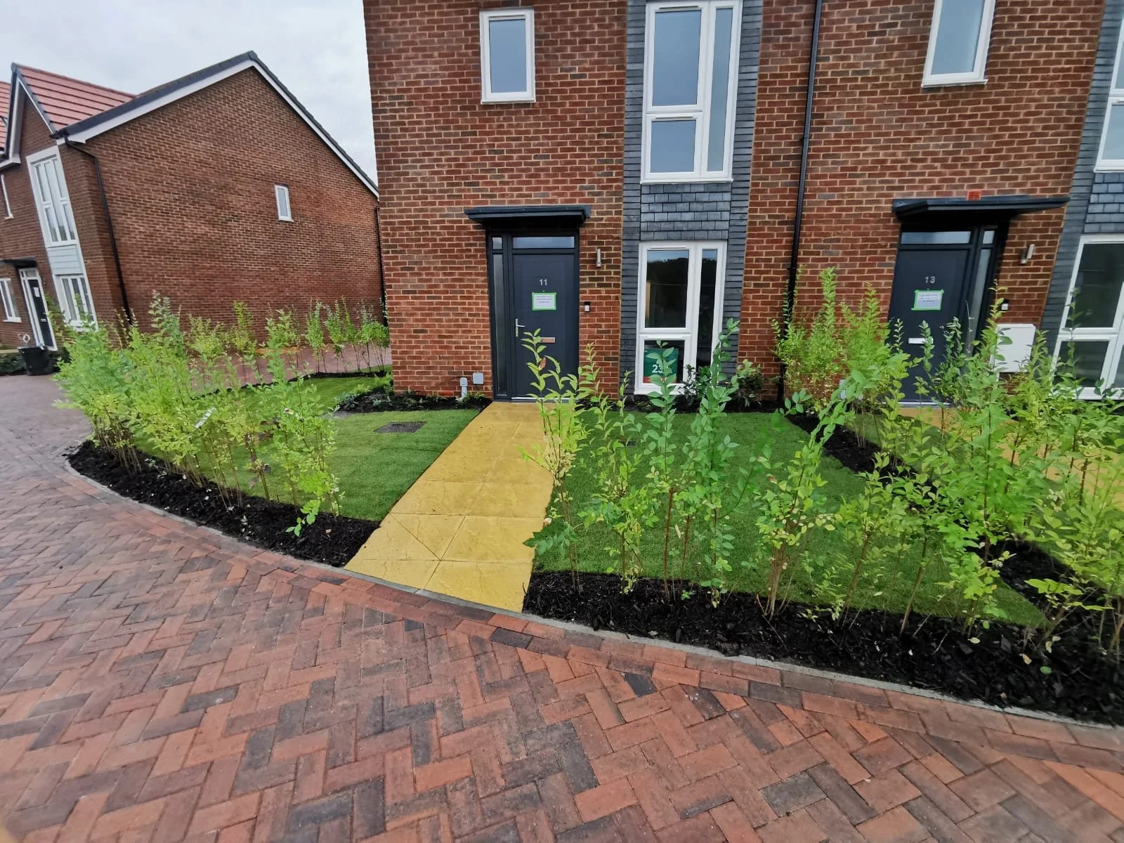 Brick residential building with black front doors, small garden with young bushes, paved sidewalk, and lawn.