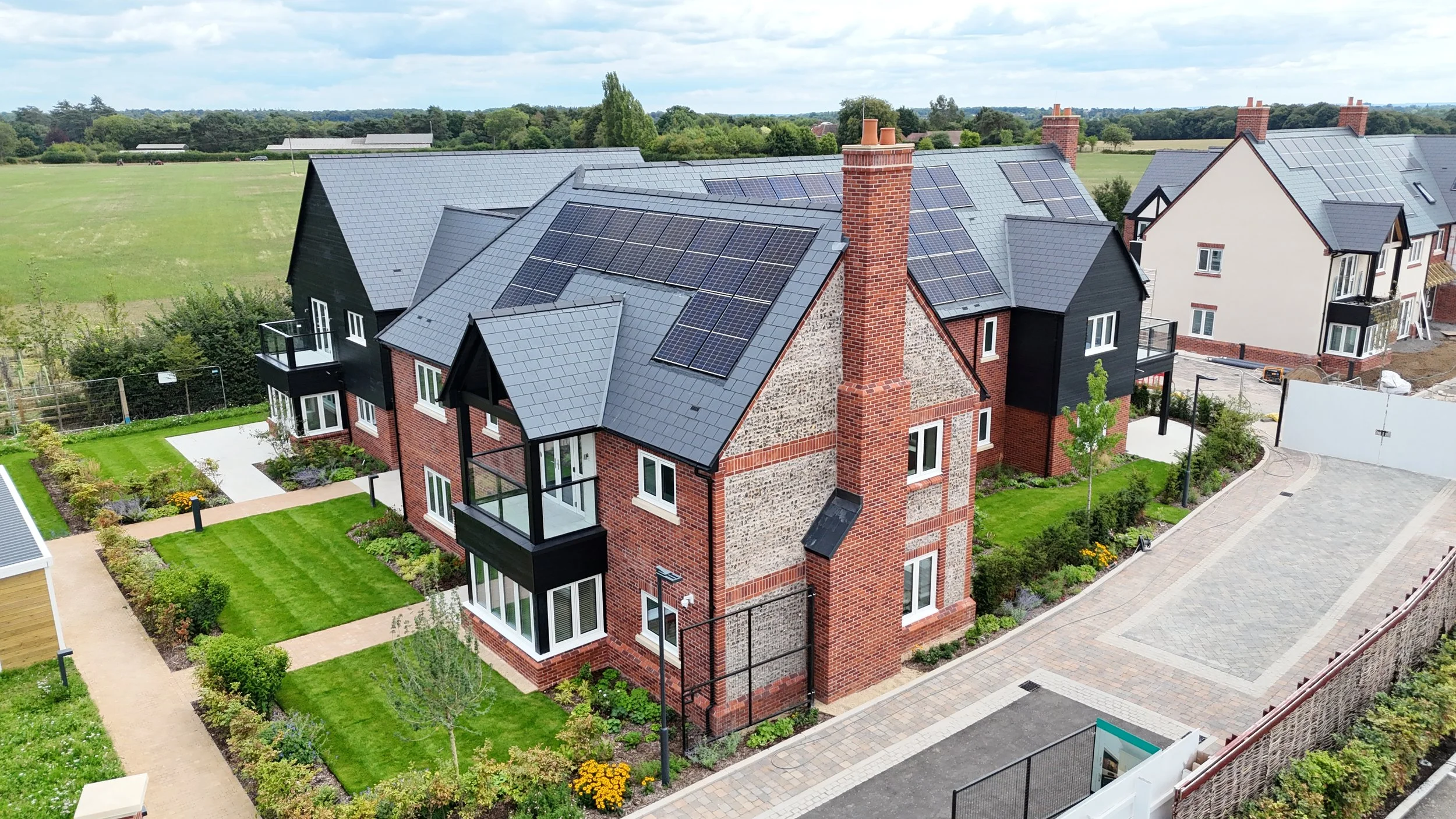 A modern house with a black and red brick exterior, solar panels on the roof, and a well-maintained garden with green grass and bushes.