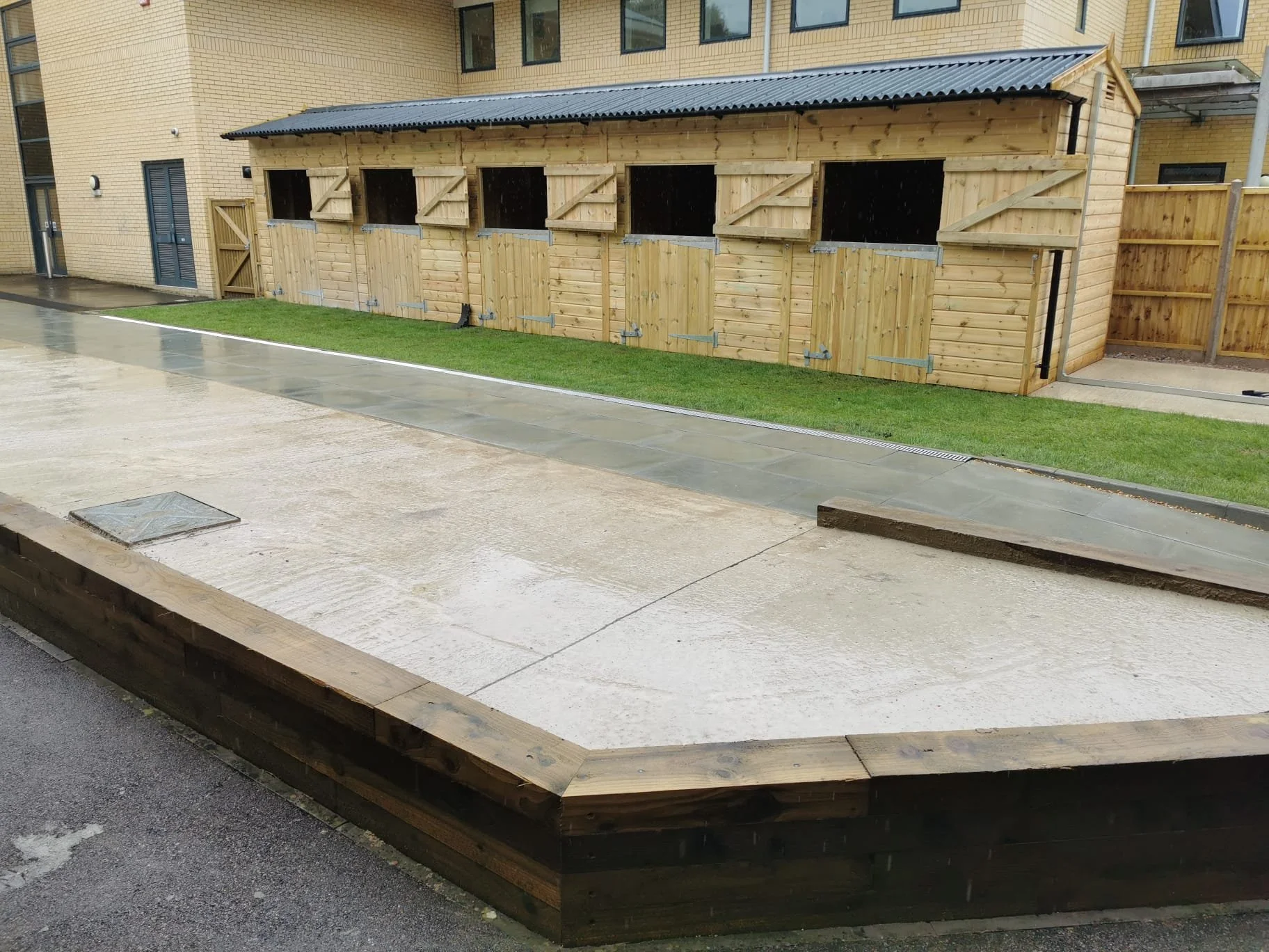 Newly built wooden storage units with multiple compartments, situated on a wet concrete area next to a grassy patch and brick residential building.