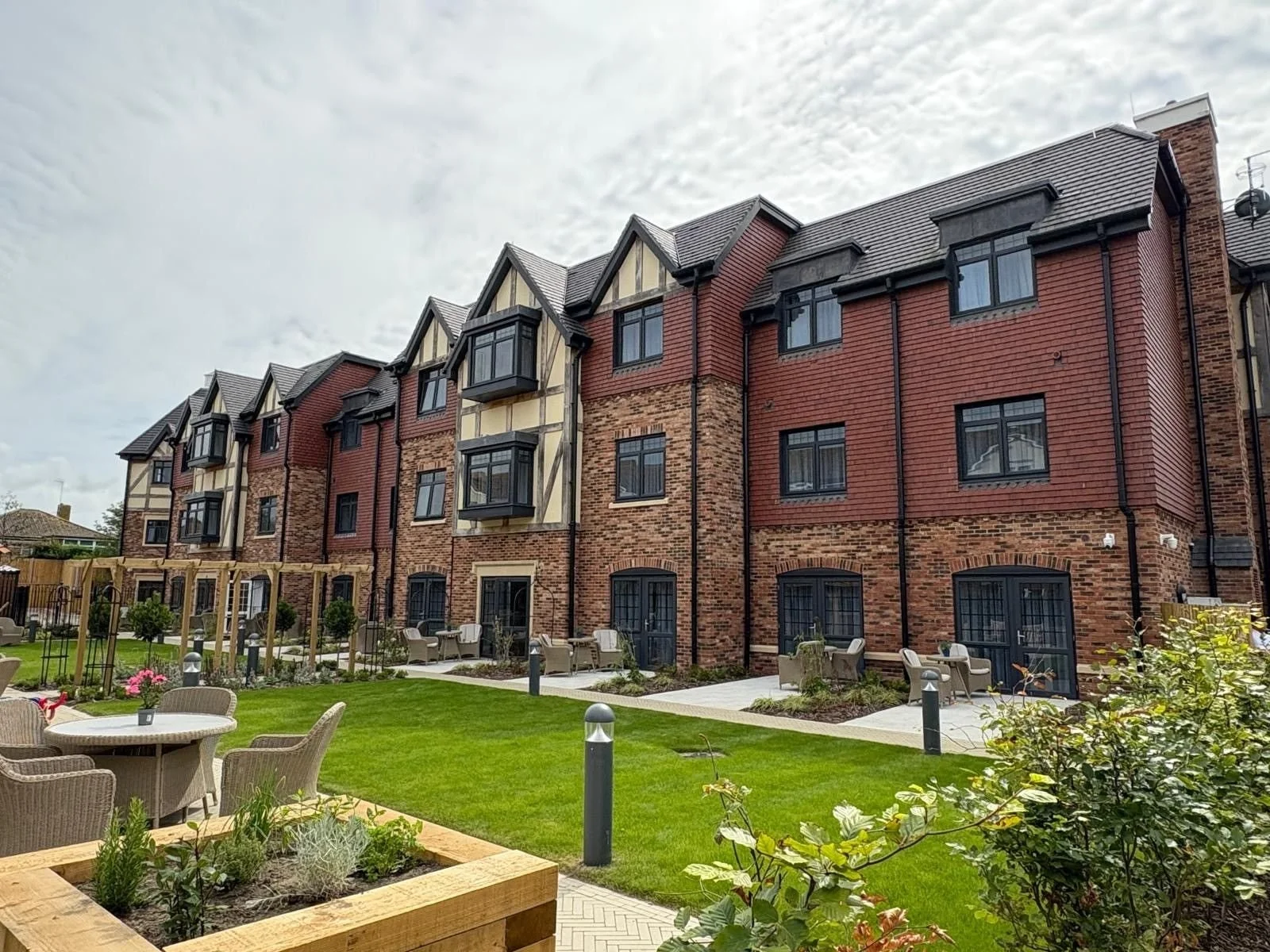 A multi-story residential building with brick and Tudor-style architecture, featuring bay windows and black window frames, overlooking a landscaped courtyard with grassy areas, patio seating, small trees, and garden beds.