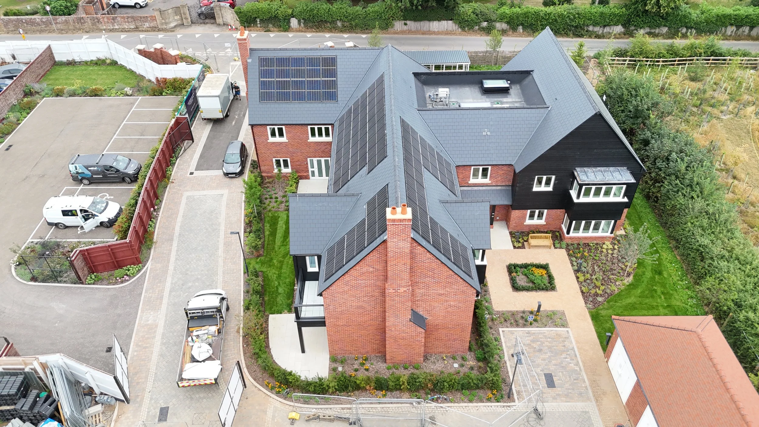 Aerial view of a modern residential building with solar panels on the roof, surrounded by landscaped gardens, parking areas, and nearby structures.