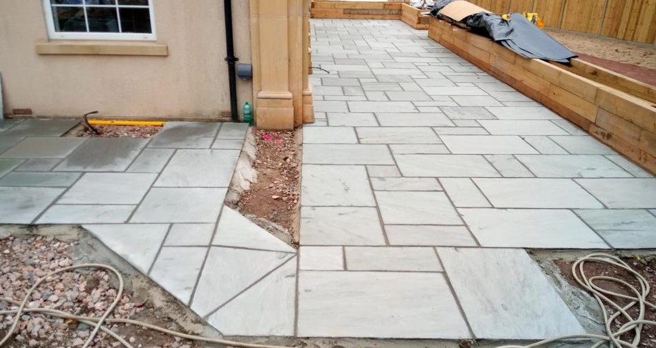 Newly installed paver patio with a mix of large rectangular and irregularly shaped stones, partially bordered with wood, next to a house with a window and a pile of materials covered with a tarp.