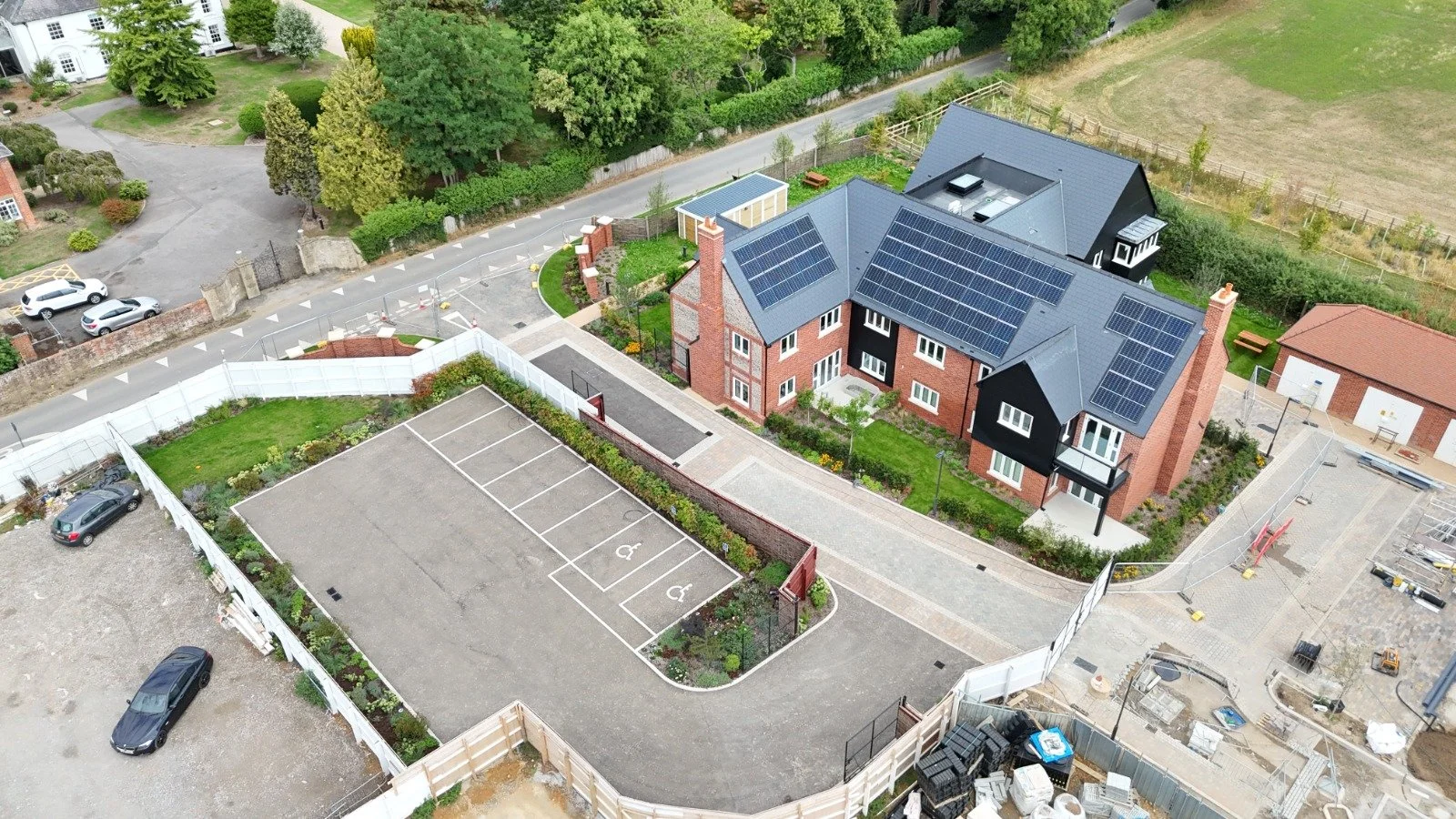 An aerial view of a residential area showing a house with solar panels on the roof, a parking lot with designated handicapped spaces, and surrounding greenery and other houses.