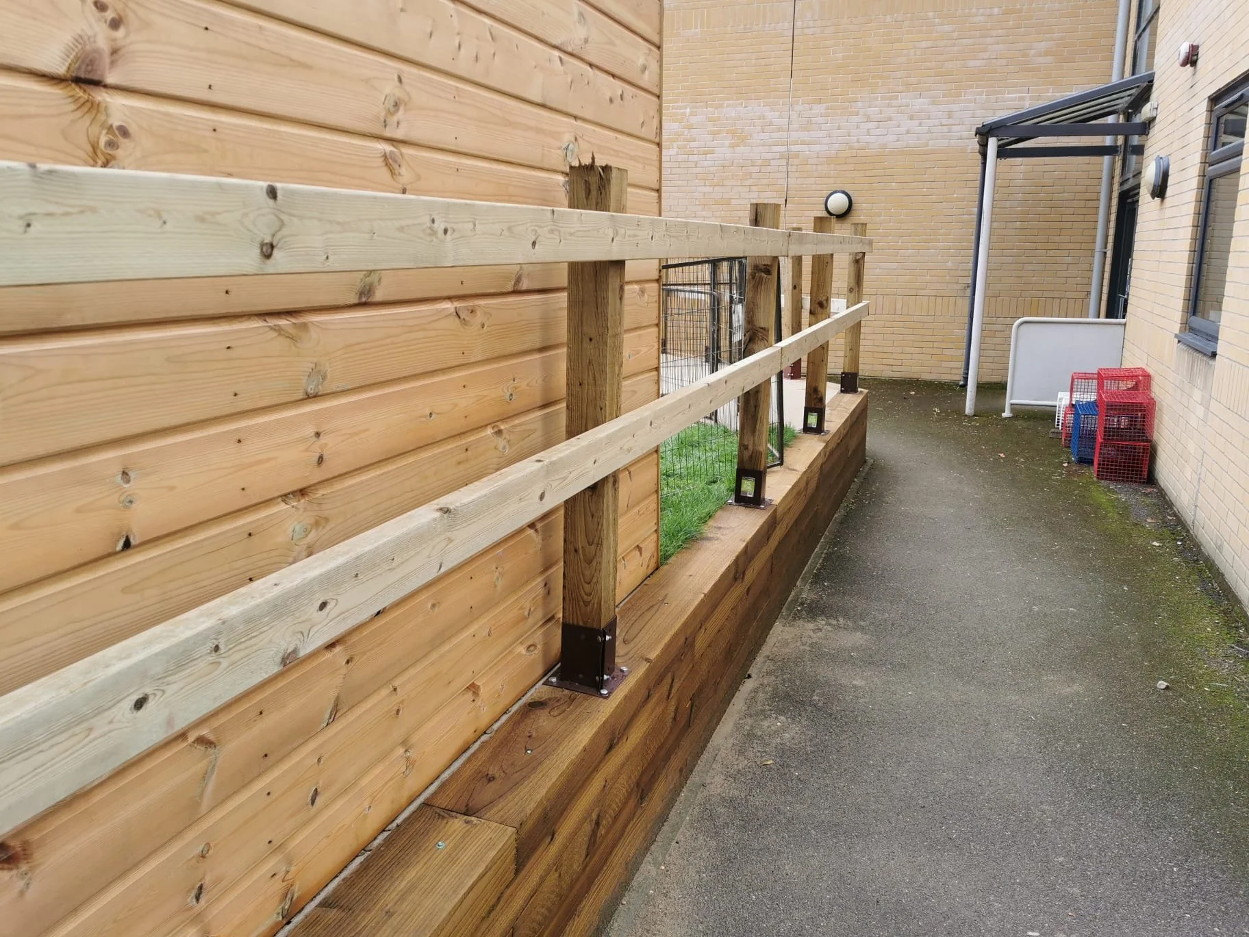 A wooden railing installed along a building exterior with a green grass patch behind using metal brackets, next to a brick wall, with a flat walkway on the other side, and some stacked red and blue crates in the background.