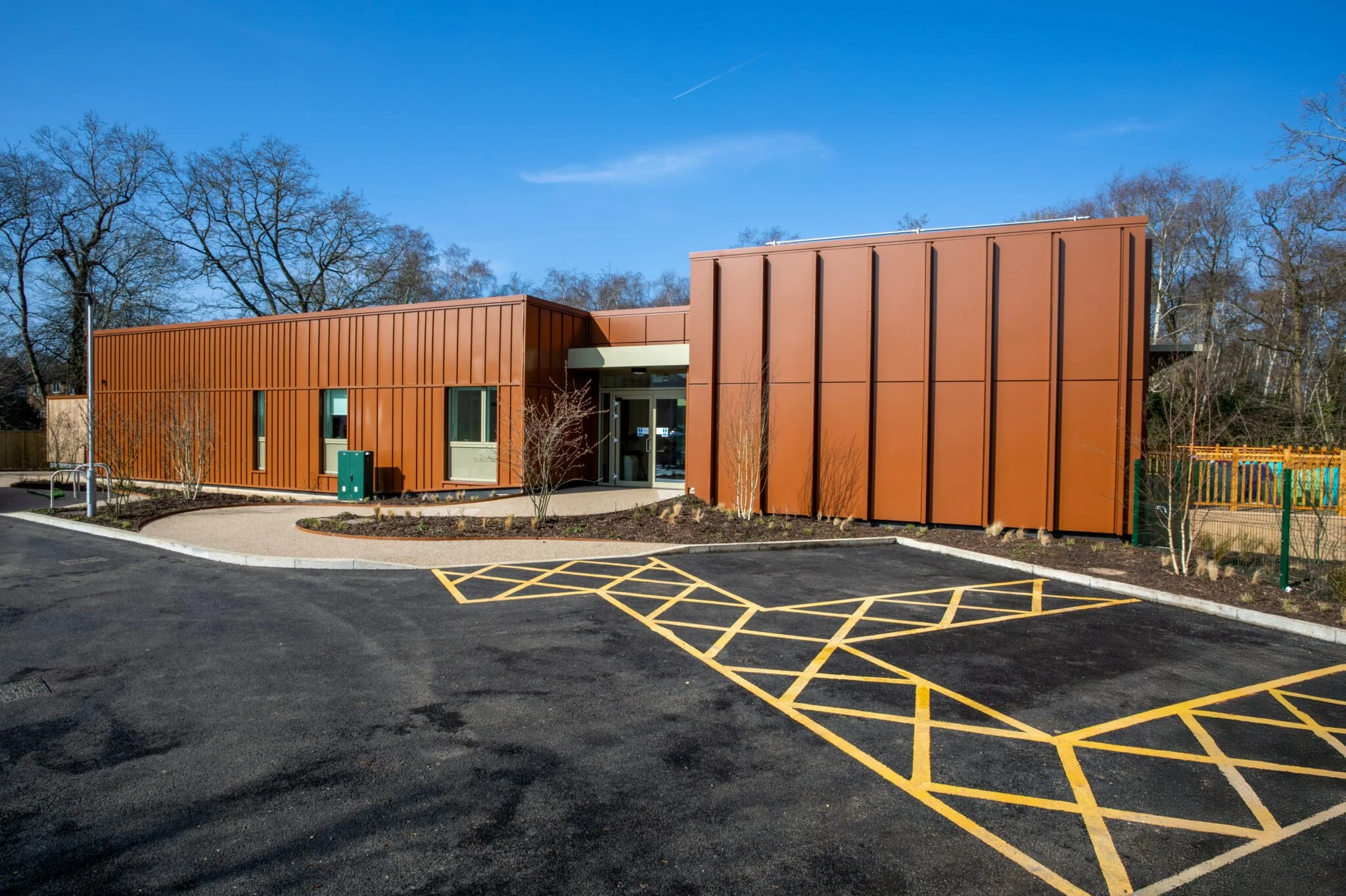 Modern single-story building with a rust-colored exterior, large windows, and a small landscaped area with young trees in front, under a clear blue sky.