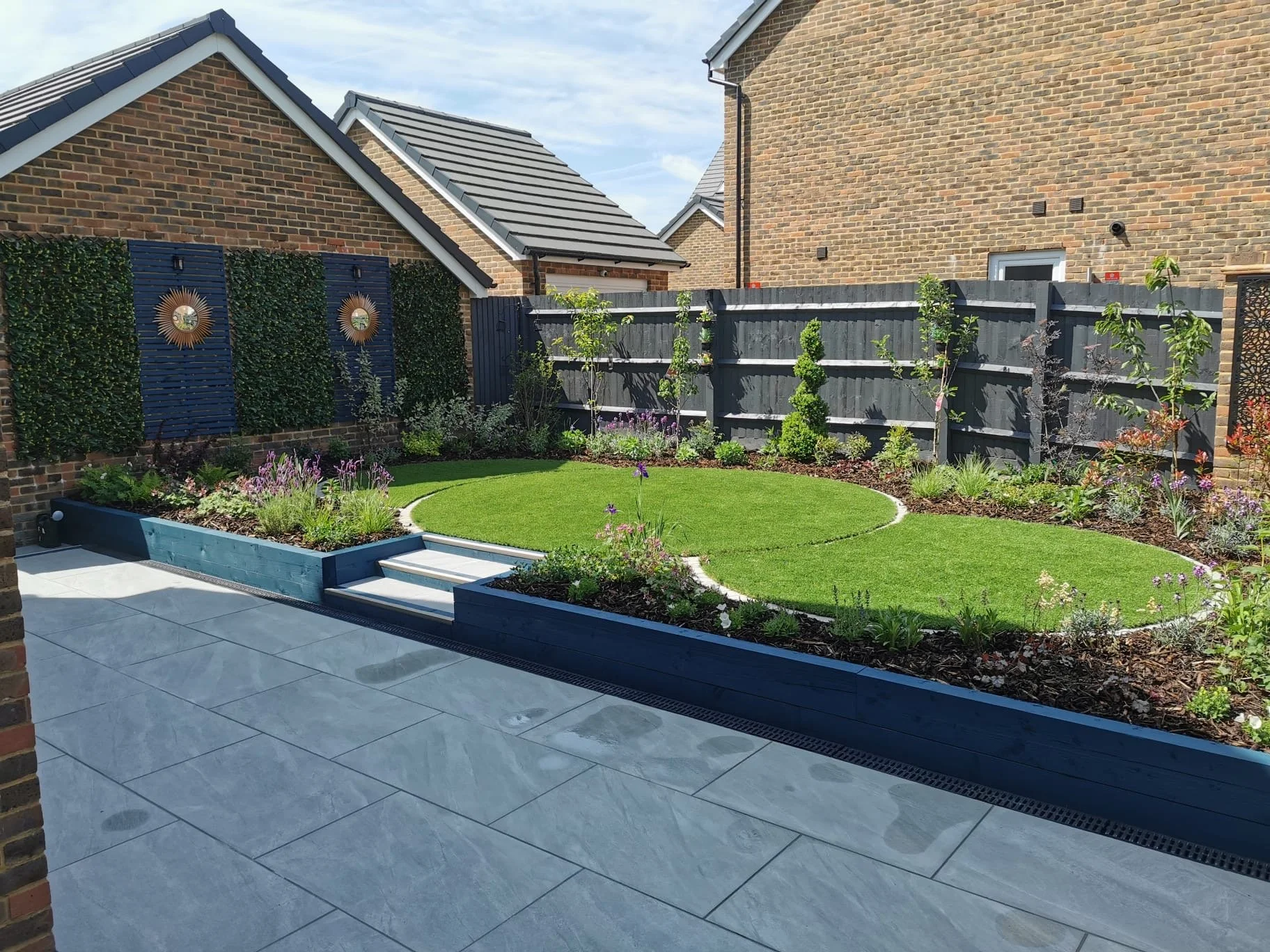 Modern backyard garden with a circular lawn, flower beds, patio, and a wooden fence with solar wind turbines for decoration.