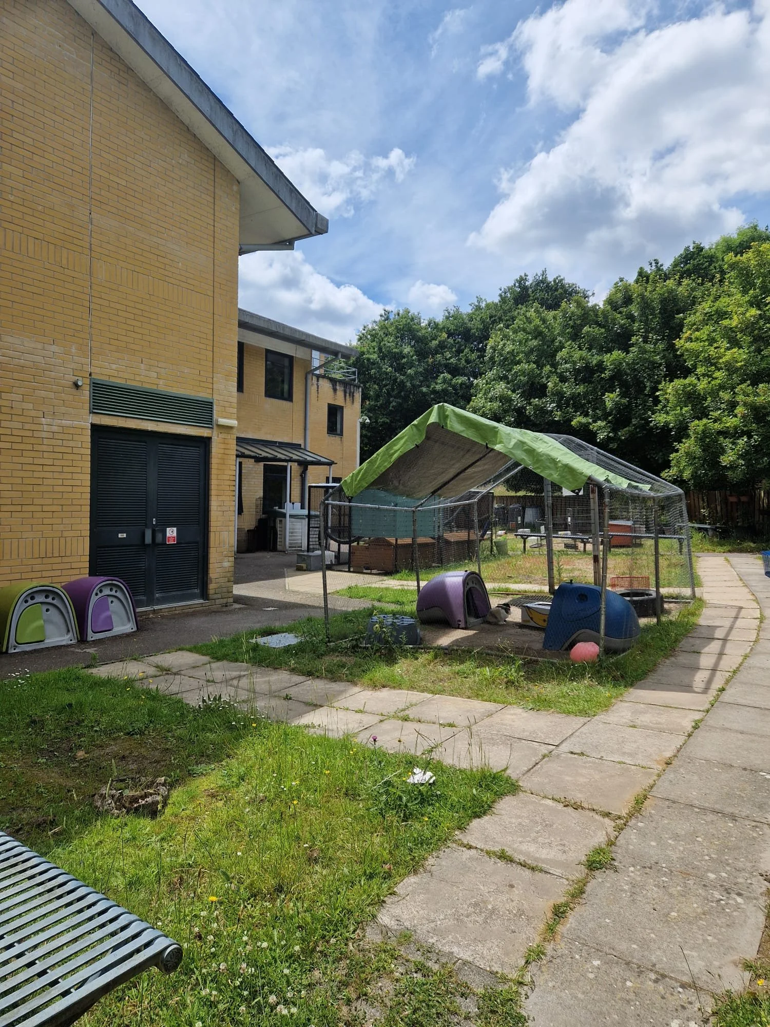 Small animal enclosure with a green canopy and colorful plastic houses, surrounded by a sidewalk and grassy area, on a sunny day with some clouds.