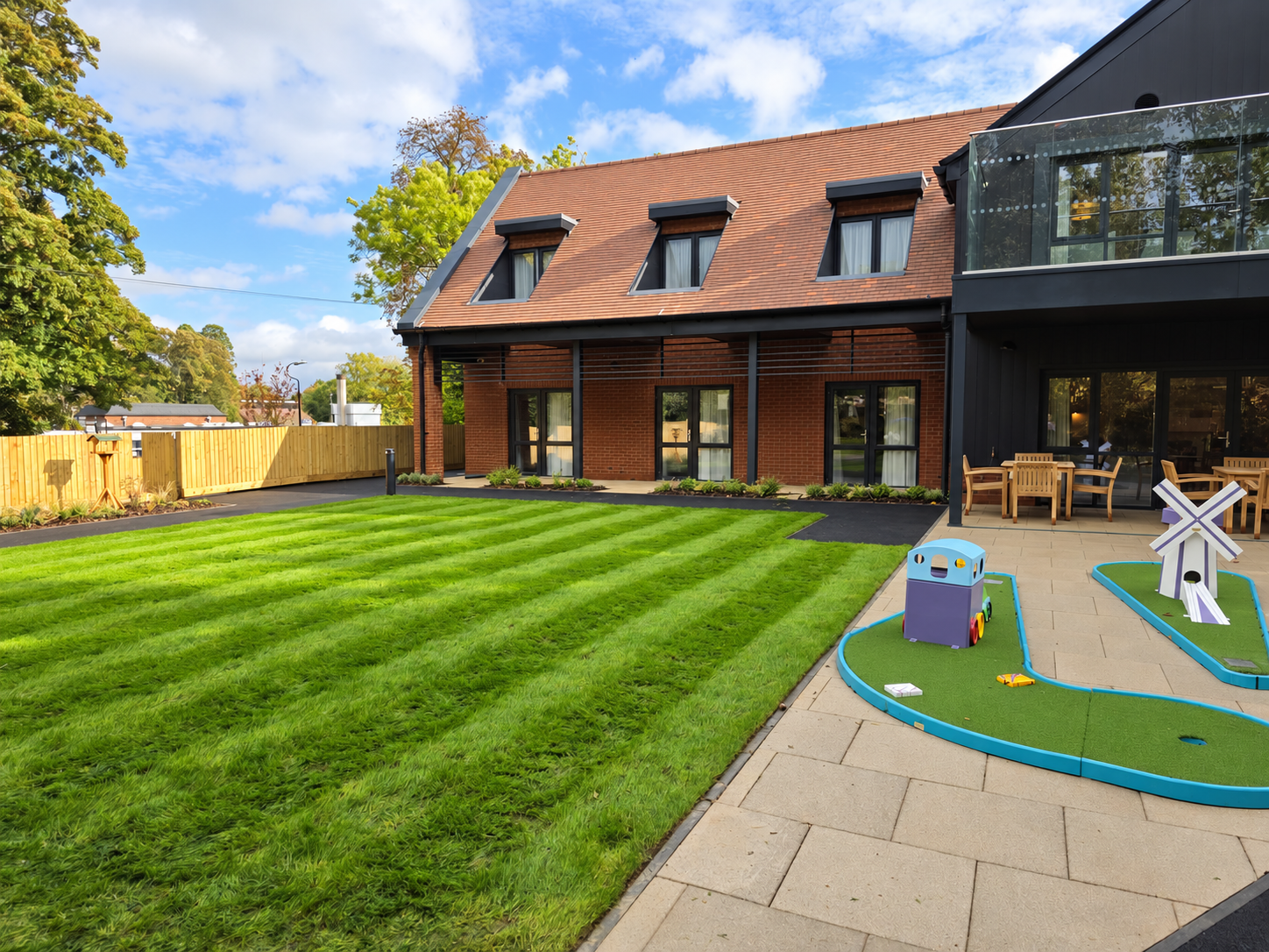 Modern backyard with freshly cut grassy lawn, patio area with outdoor furniture, and children's play area with a small toy train and windmill gnome.