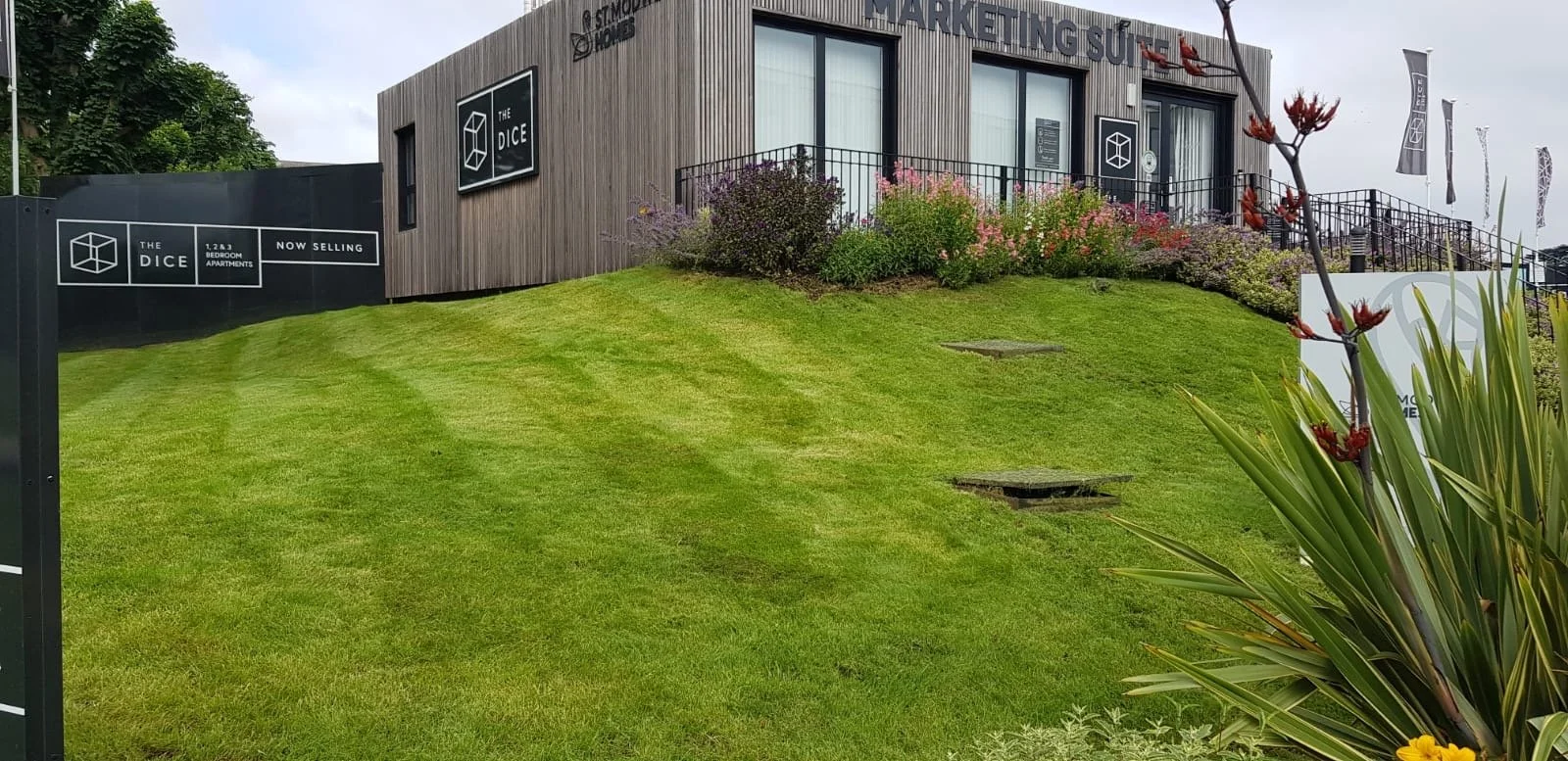 Exterior view of a modern building with signs for 'The Dice' and an outdoor marketing suite, surrounded by green grass, flowering bushes, and a few plants.