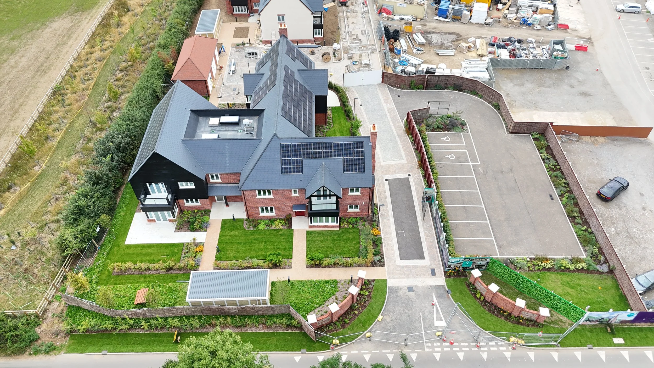 An aerial view of a large residential building with solar panels on its roof, surrounded by landscaped gardens, a parking lot, and construction area with equipment and materials.