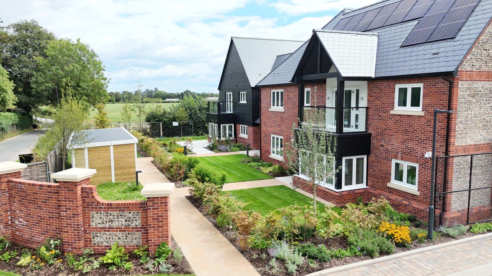 Modern brick residential building with black and brick sections, solar panels on the roof, and landscaped garden with green grass, shrubs, and trees.