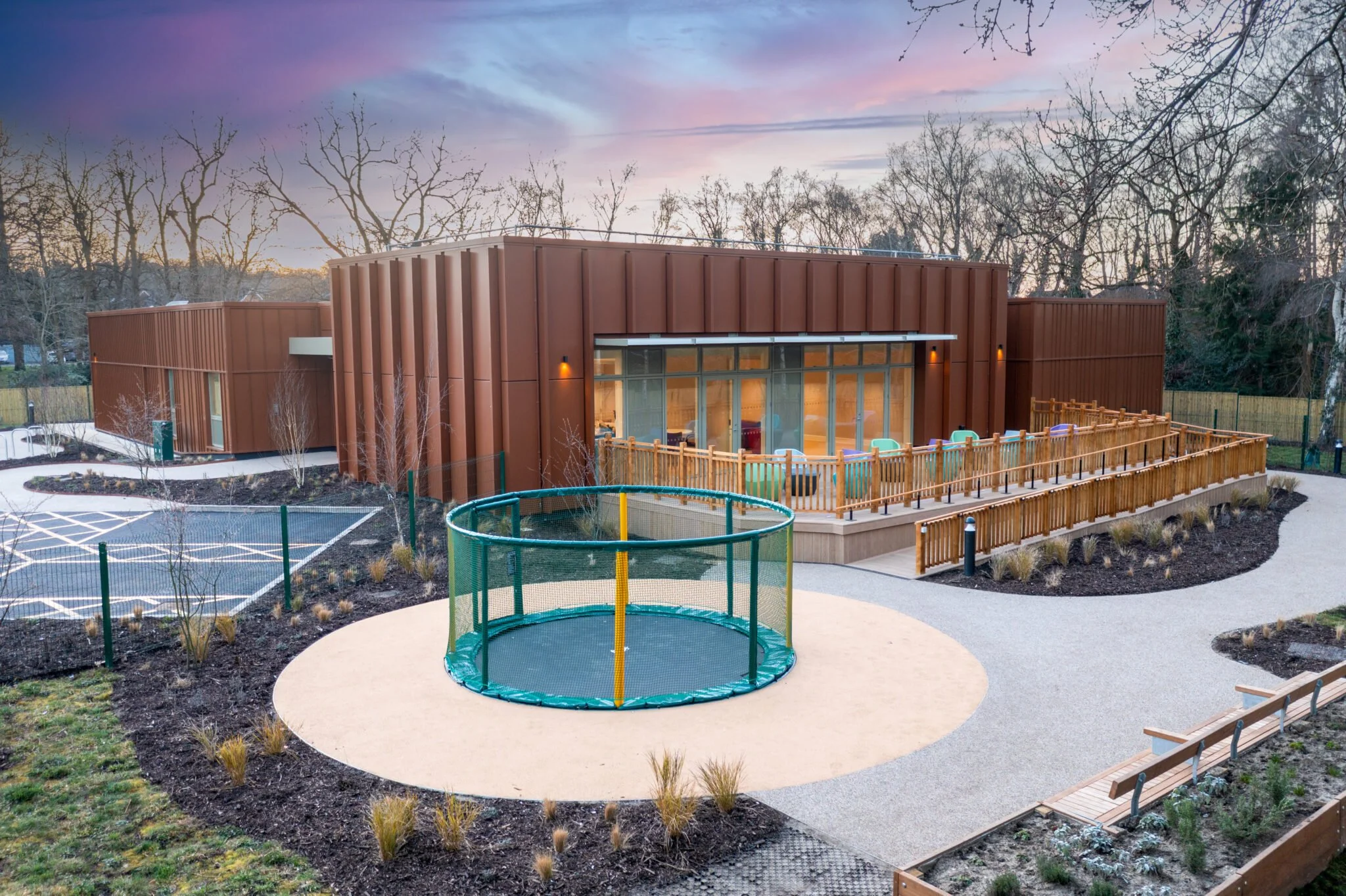 Modern brown building with large glass windows, surrounded by a landscaped yard with a gravel pathway, a circular trampoline, and outdoor seating area, during sunset.