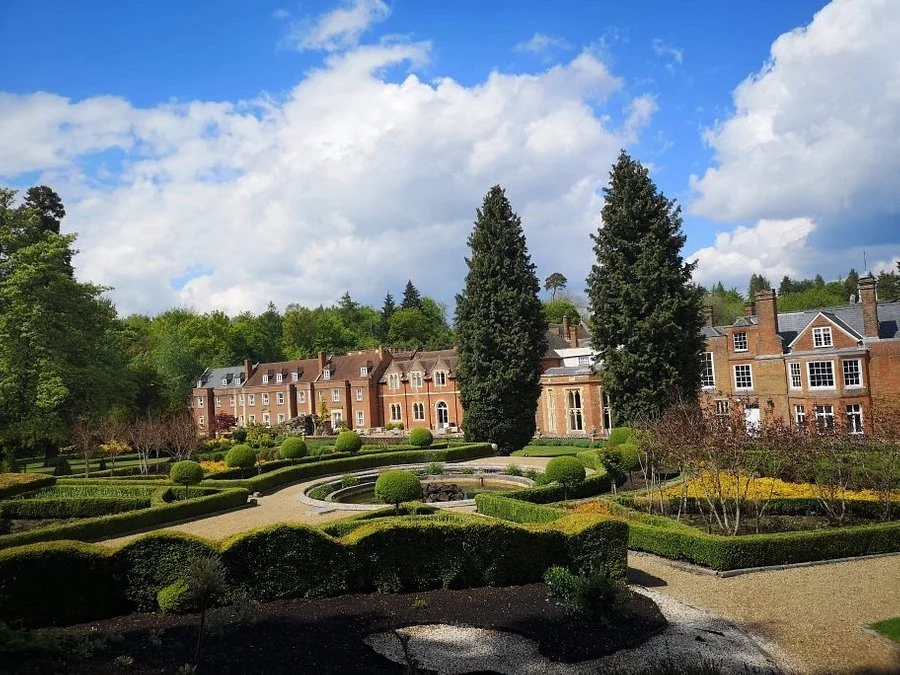 A historic brick mansion surrounded by a landscaped garden with trimmed bushes, tall trees, and walking paths under a partly cloudy blue sky.