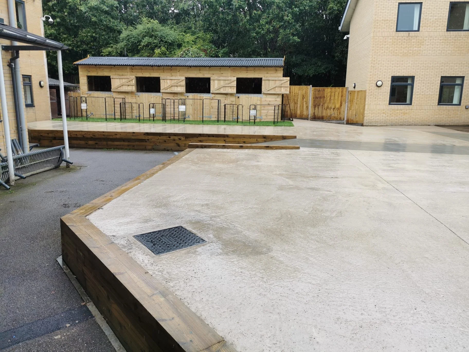 A playground sandbox with wooden borders, a drain, and a wooden building in the background surrounded by a metal fence, with a brick apartment building and trees behind.
