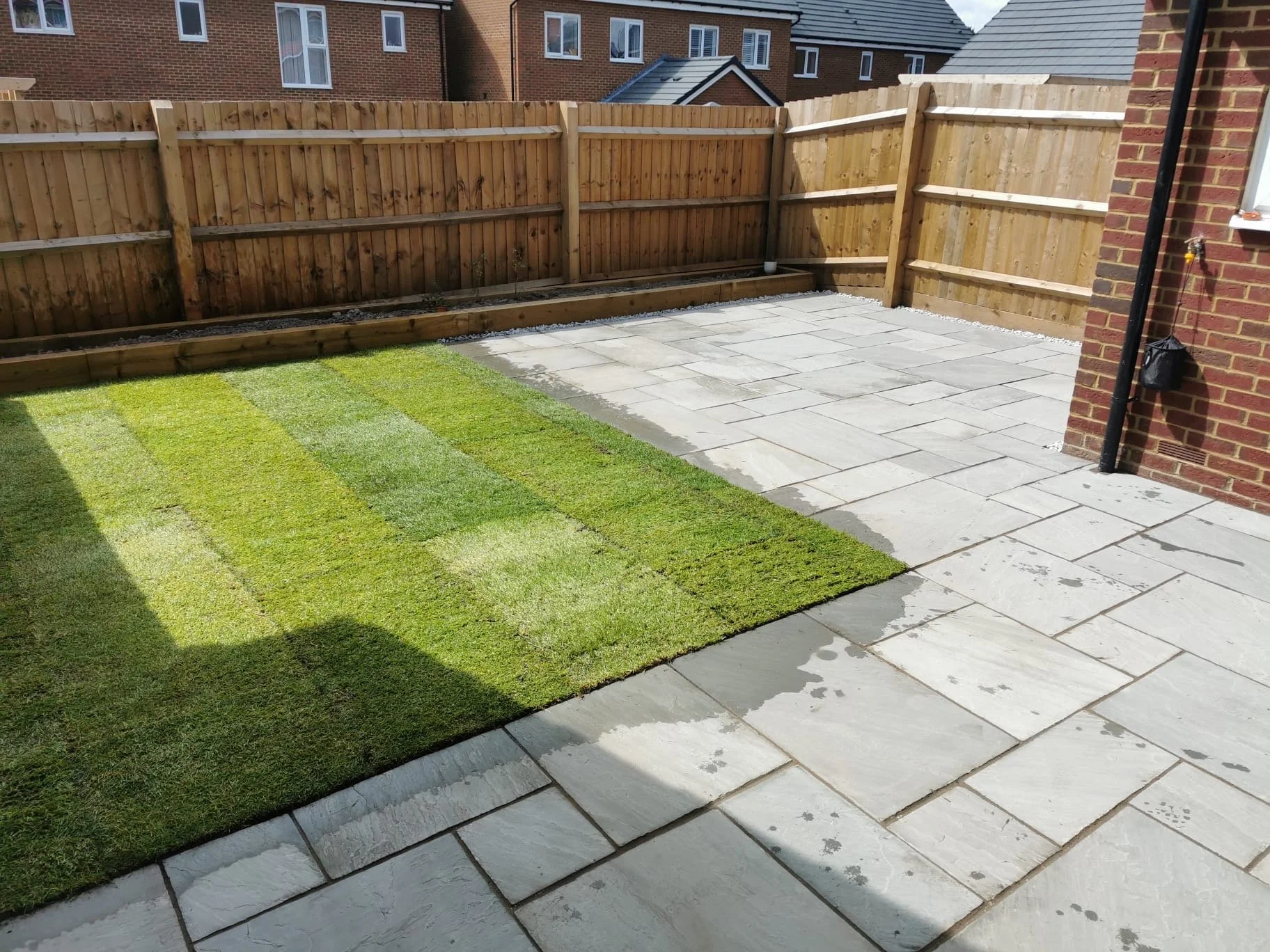 Backyard patio with green grass and gray stone paving, enclosed by a wooden fence and brick house exterior.