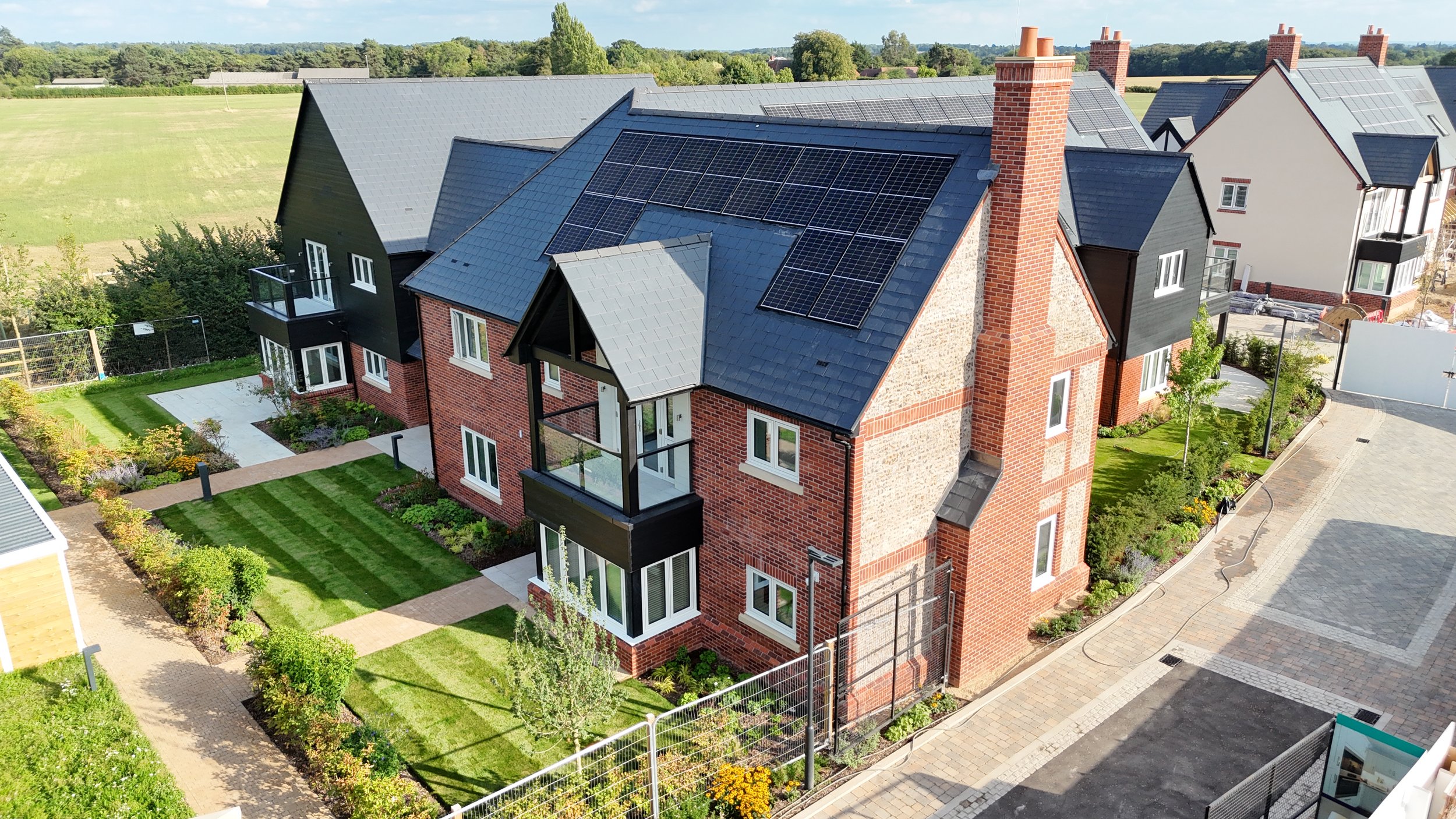 A modern multi-story residential building with solar panels on the roof and a well-maintained garden with green lawns, shrubs, and small trees, in a suburban neighborhood.
