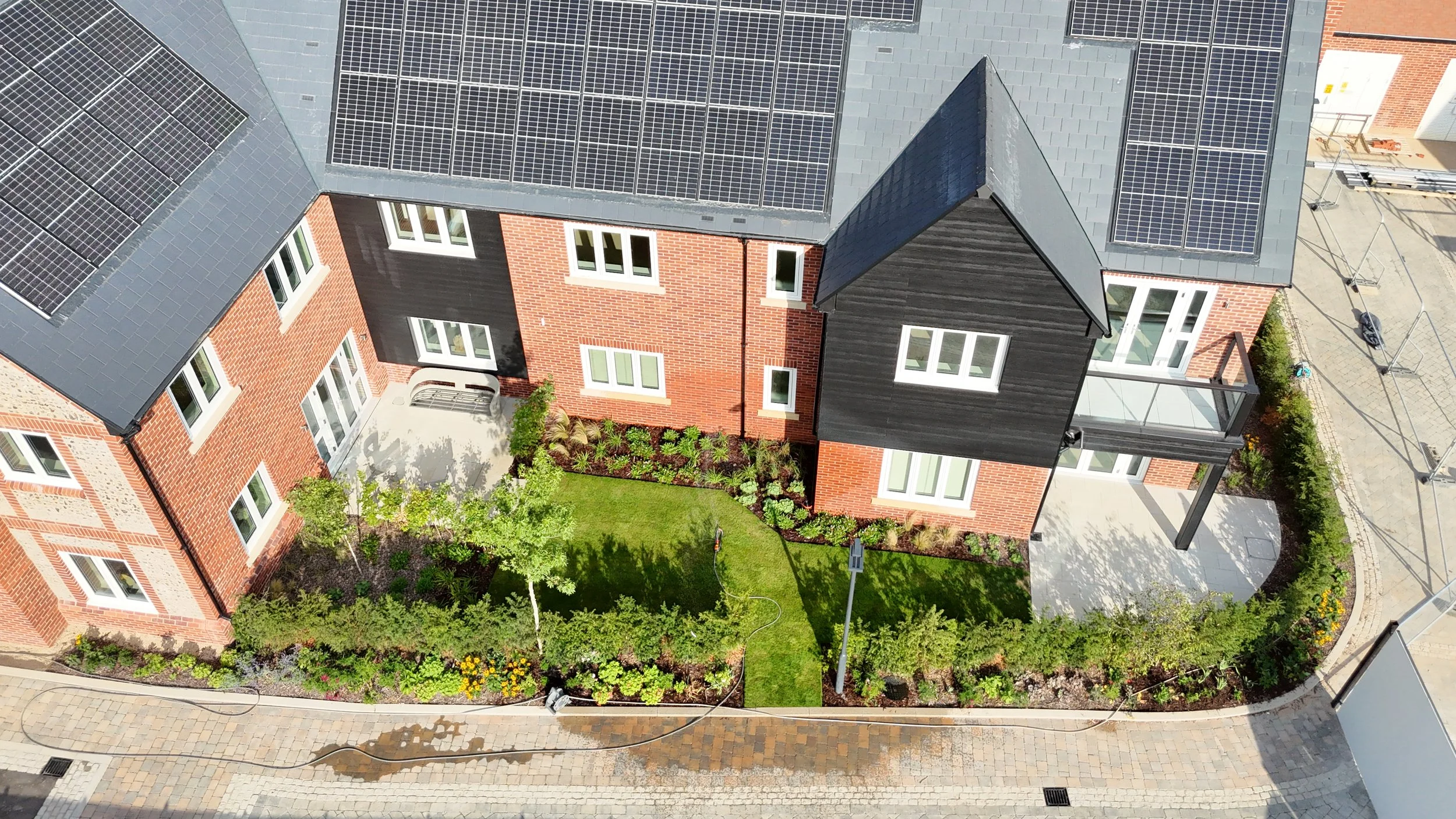 Aerial view of a modern residential building with red brick walls, dark gray roof, and solar panels. The yard features a small lawn, garden beds, and a paved patio area.