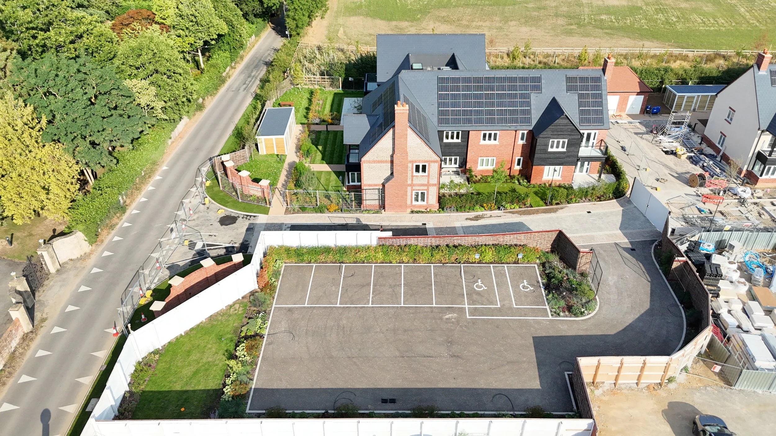 Aerial view of a residential area showing a building with solar panels on the roof, an adjacent parking lot with designated handicapped spaces, a garden, and a street lined with trees.