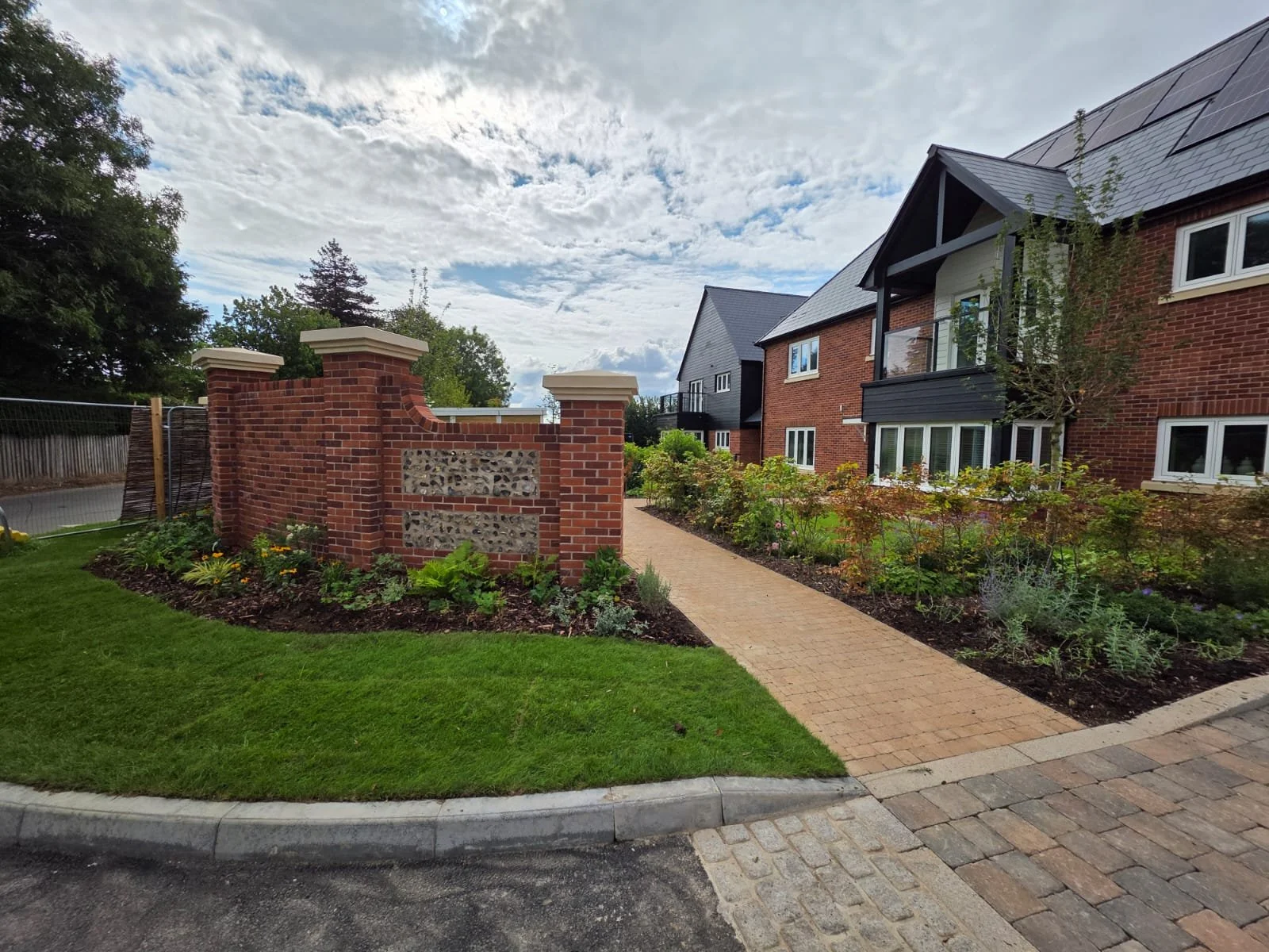 Residential apartment building with landscaped garden, brick and black exterior, paved walkway, and a brick wall entrance, under a cloudy sky.
