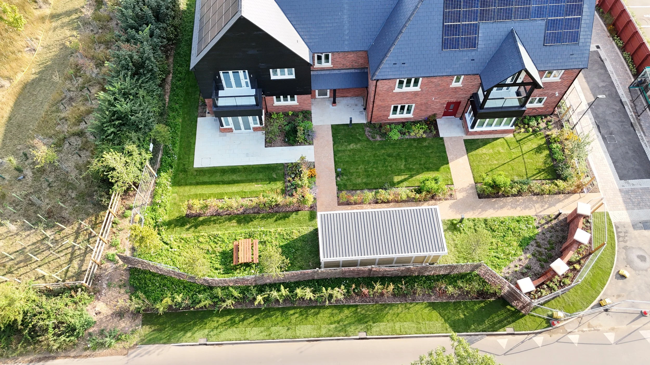 Aerial view of a backyard with green lawns, garden beds, a small shed, and multiple walkways, surrounded by a fence with neighboring buildings visible.