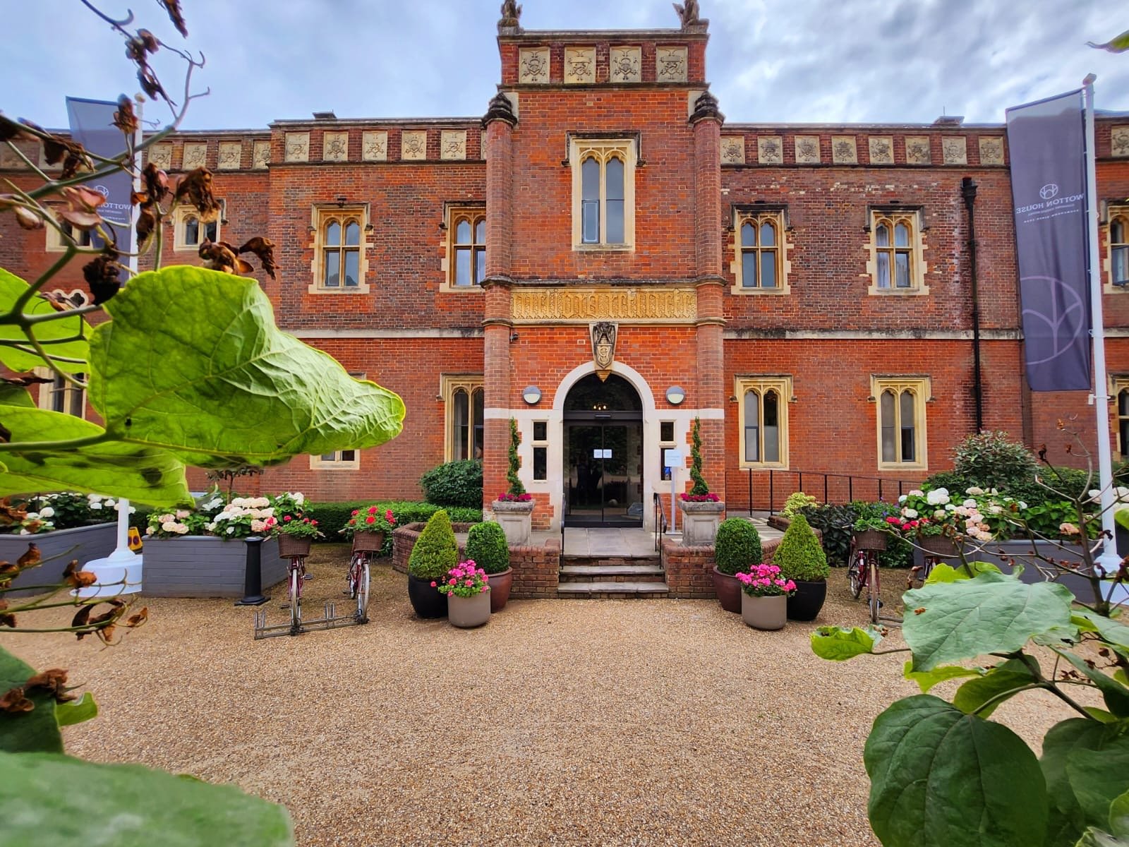 Front view of a red brick building with Gothic architecture, arched doorway, and stained glass windows, decorated with potted plants and flowers, with banners on either side.
