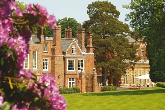 A large historic brick mansion with multiple chimneys surrounded by a well-manicured lawn and tall trees, with pink flowers in the foreground.