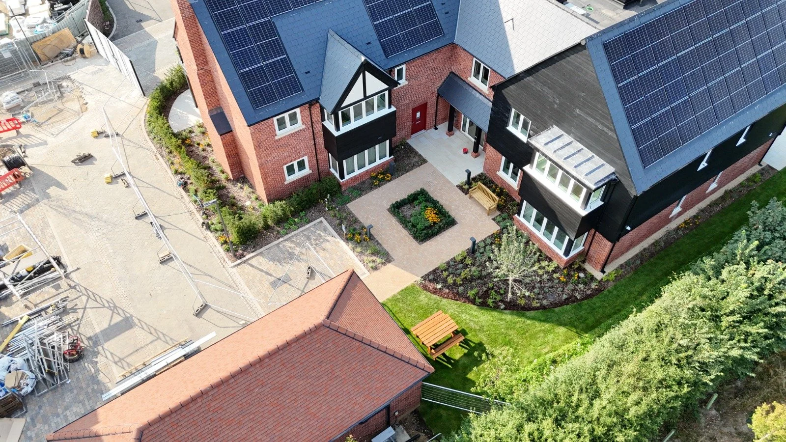 Aerial view of a residential house with solar panels on the roof, a landscaped front yard with a small garden, a bench, and paved pathways. Adjacent area shows construction materials and equipment.
