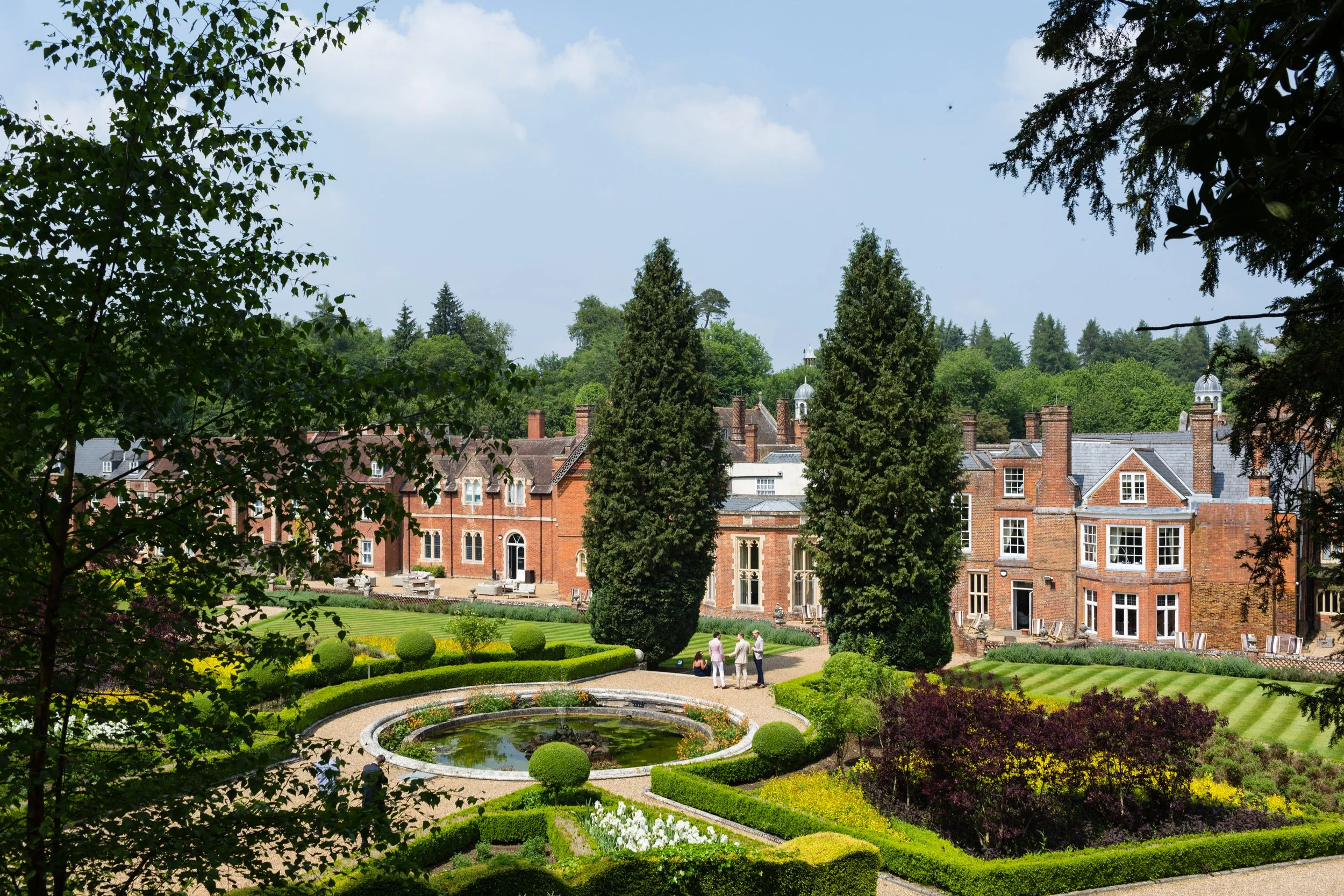 A scenic view of a historic brick mansion surrounded by a beautifully maintained garden with a pond, trimmed bushes, and tall trees under a partly cloudy sky.