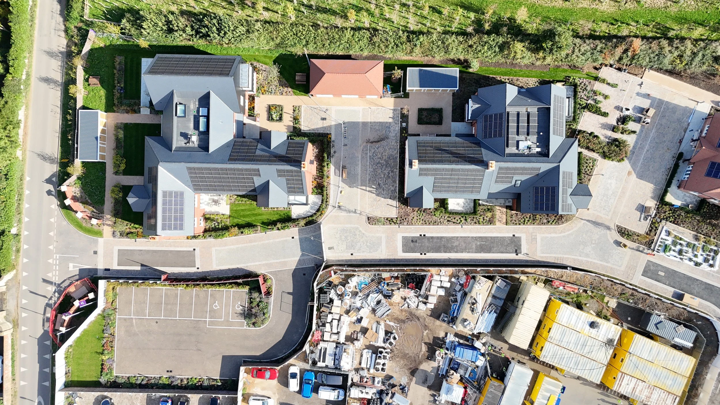 An aerial view of a residential area with three buildings featuring solar panels on their roofs, surrounded by well-maintained gardens, parking lots, and a construction site with scattered materials and vehicles.
