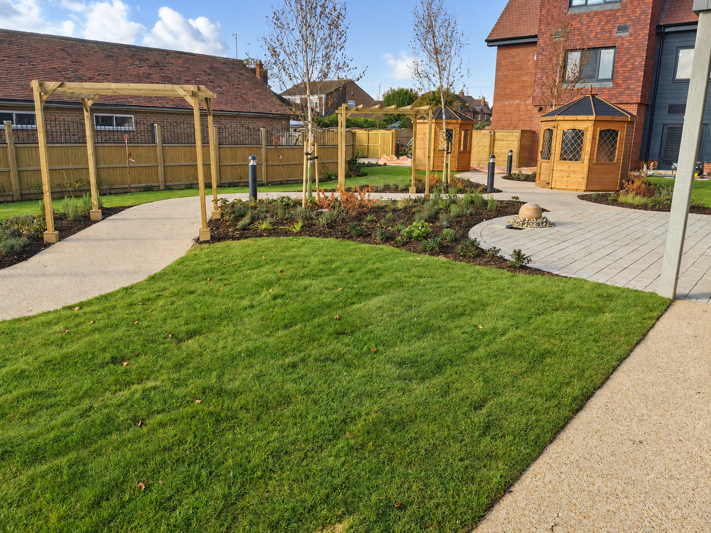 A landscaped backyard with a green lawn, winding concrete and stone pathways, small wooden garden sheds, and newly planted trees, enclosed by a wooden fence.