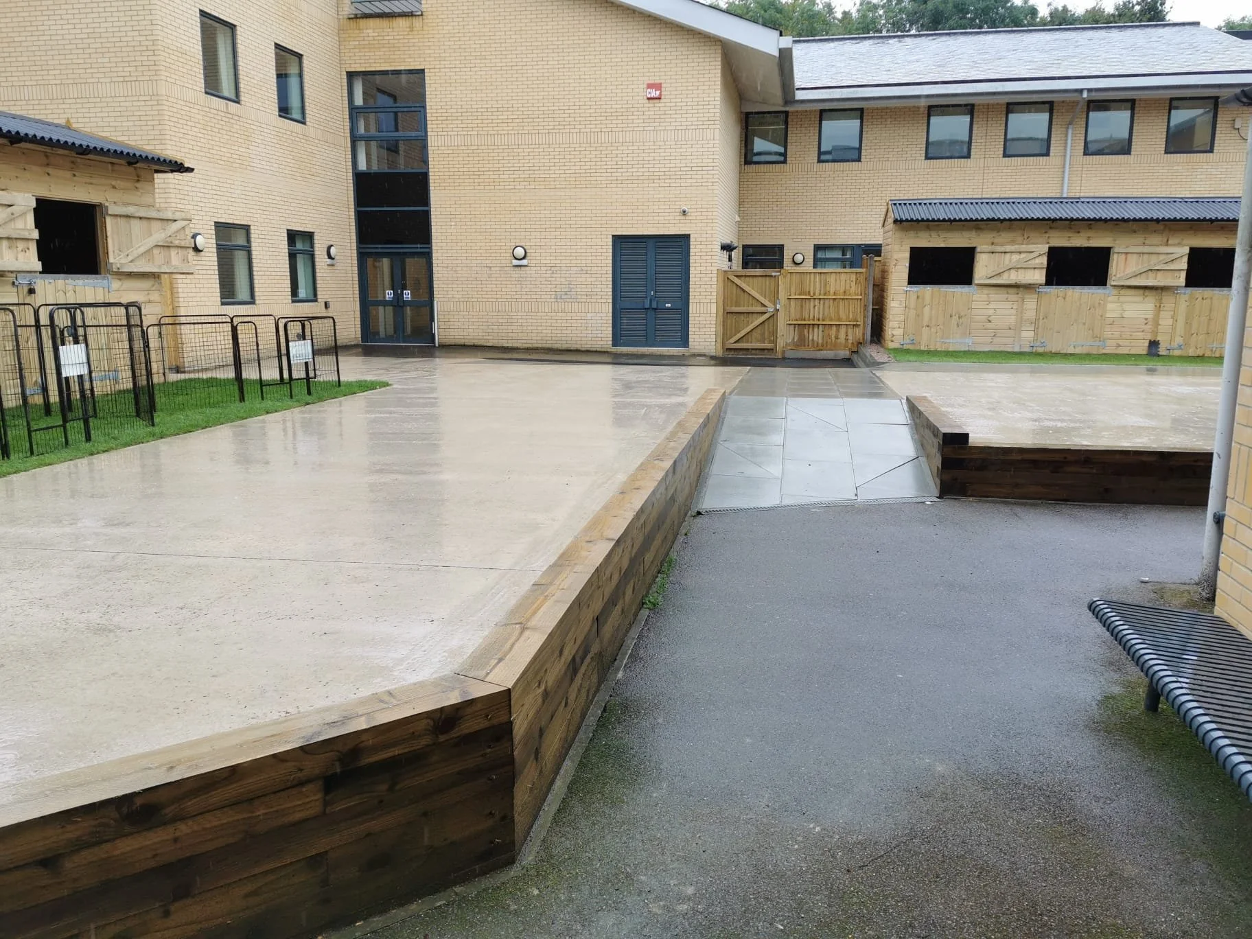 Rainy courtyard with wet concrete and wooden fencing, building with brick walls, black and blue doors, and several small windows.