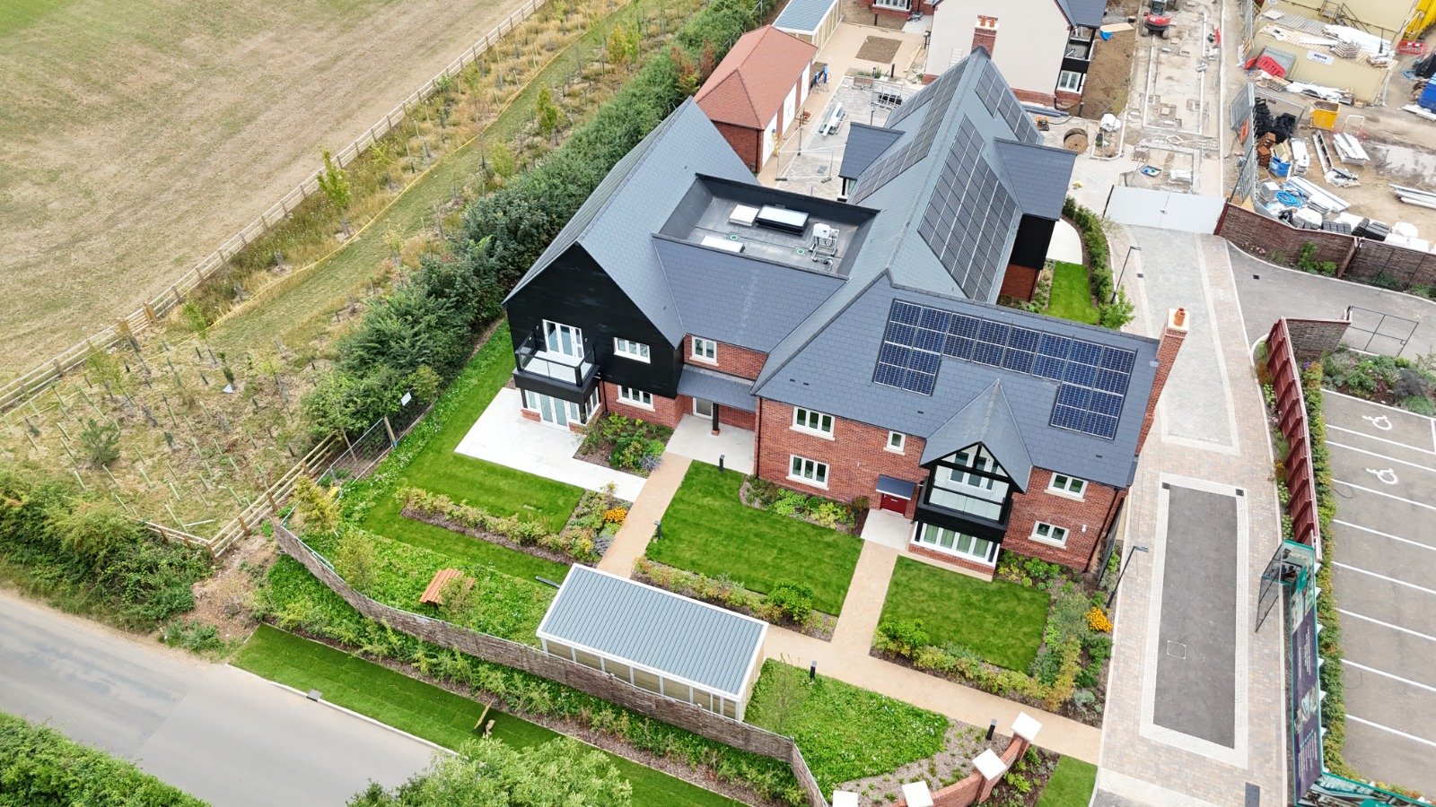 Aerial view of a modern residential house with solar panels on the roof, surrounded by landscaped garden with pathways, parking lot, and adjacent construction site.