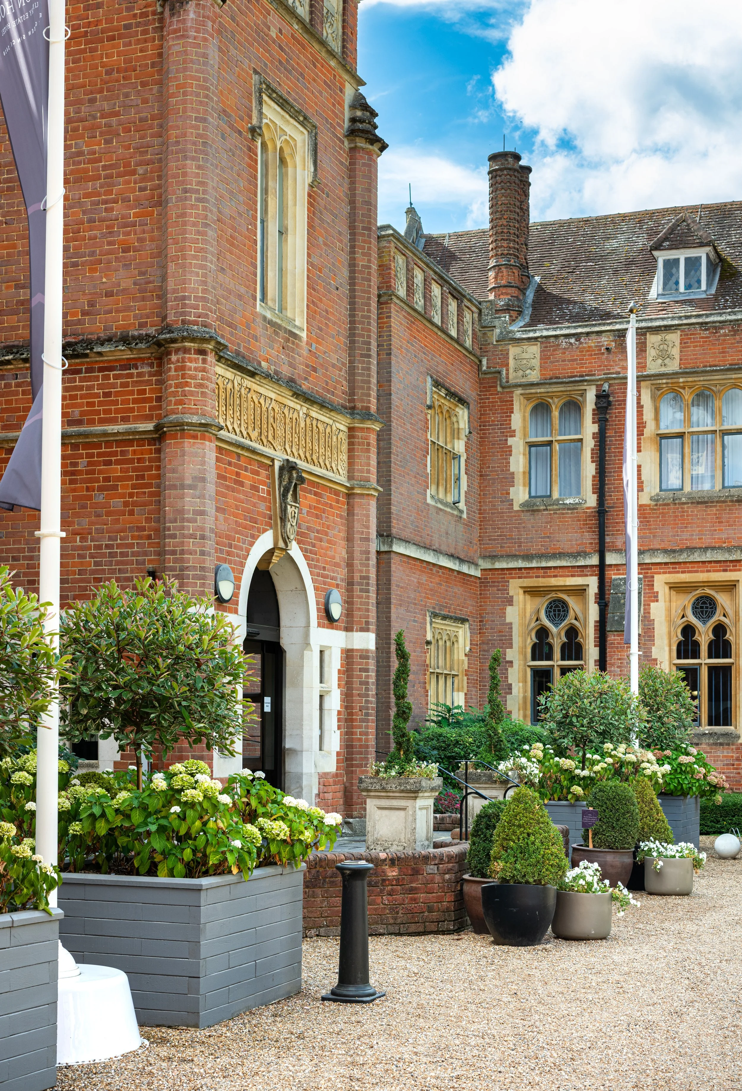 A close-up view of a historic red brick building with ornate window details, surrounded by potted plants, shrubs, and flowers in a garden area, with a gravel pathway and a blue sky with some clouds overhead.